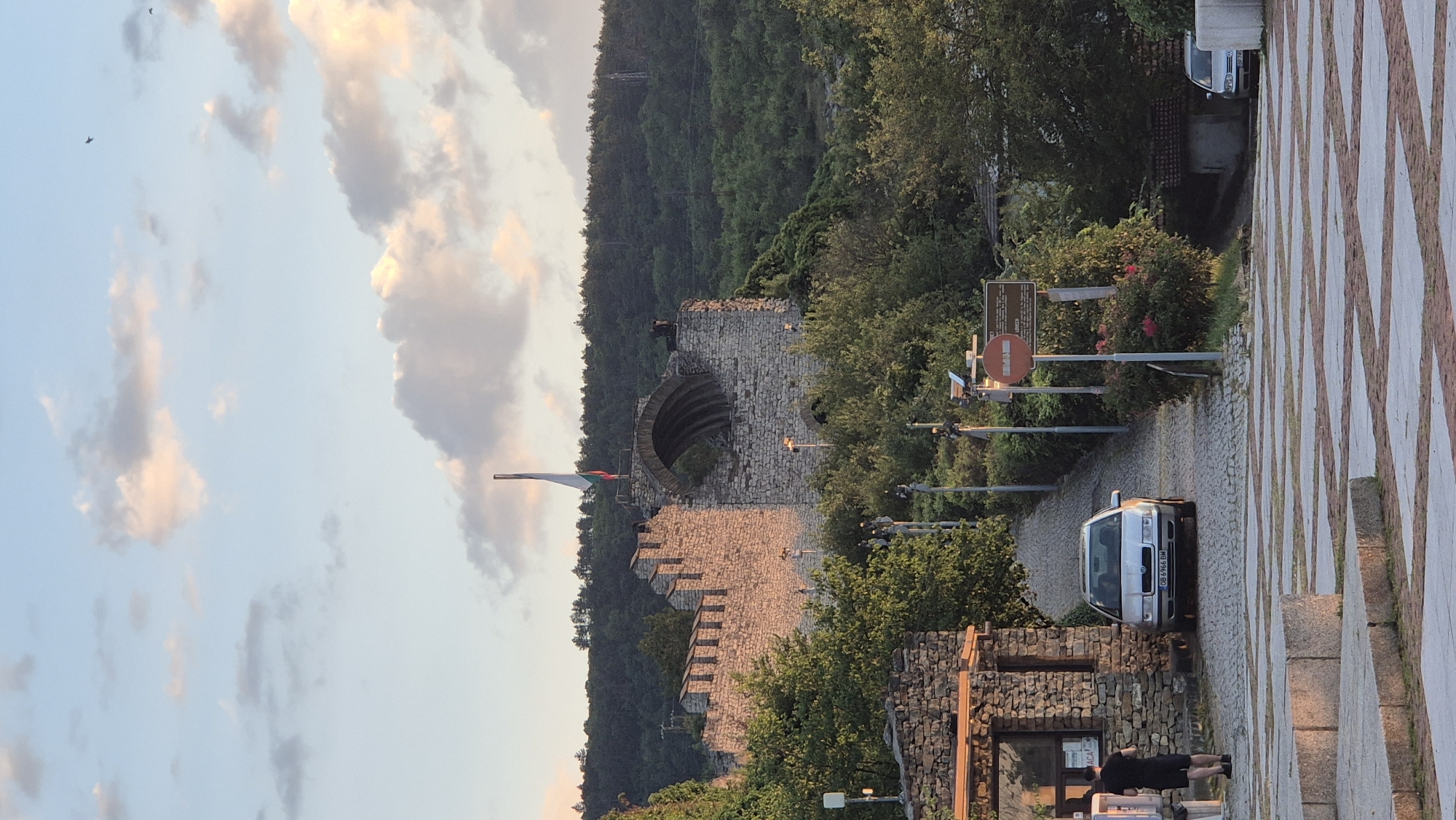 The image depicts the Tsarevets Fortress in Veliko Tarnovo, Bulgaria, at sunset. A stone wall with crenellations runs horizontally across the mid-ground, with a large arched opening in the center. A flag, appearing to be the Bulgarian flag, flies from a pole above the arch. The fortress is surrounded by lush green trees, and the sky is filled with soft, cloudy hues of pink and orange, indicative of sunset. In the foreground, a cobblestone path leads up to the fortress, with a silver sedan parked on the right side of the path. A person in a black t-shirt and shorts stands near a stone building on the left. Another car is partially visible on the far right. The overall scene conveys a sense of historical grandeur and natural beauty bathed in the warm light of dusk.