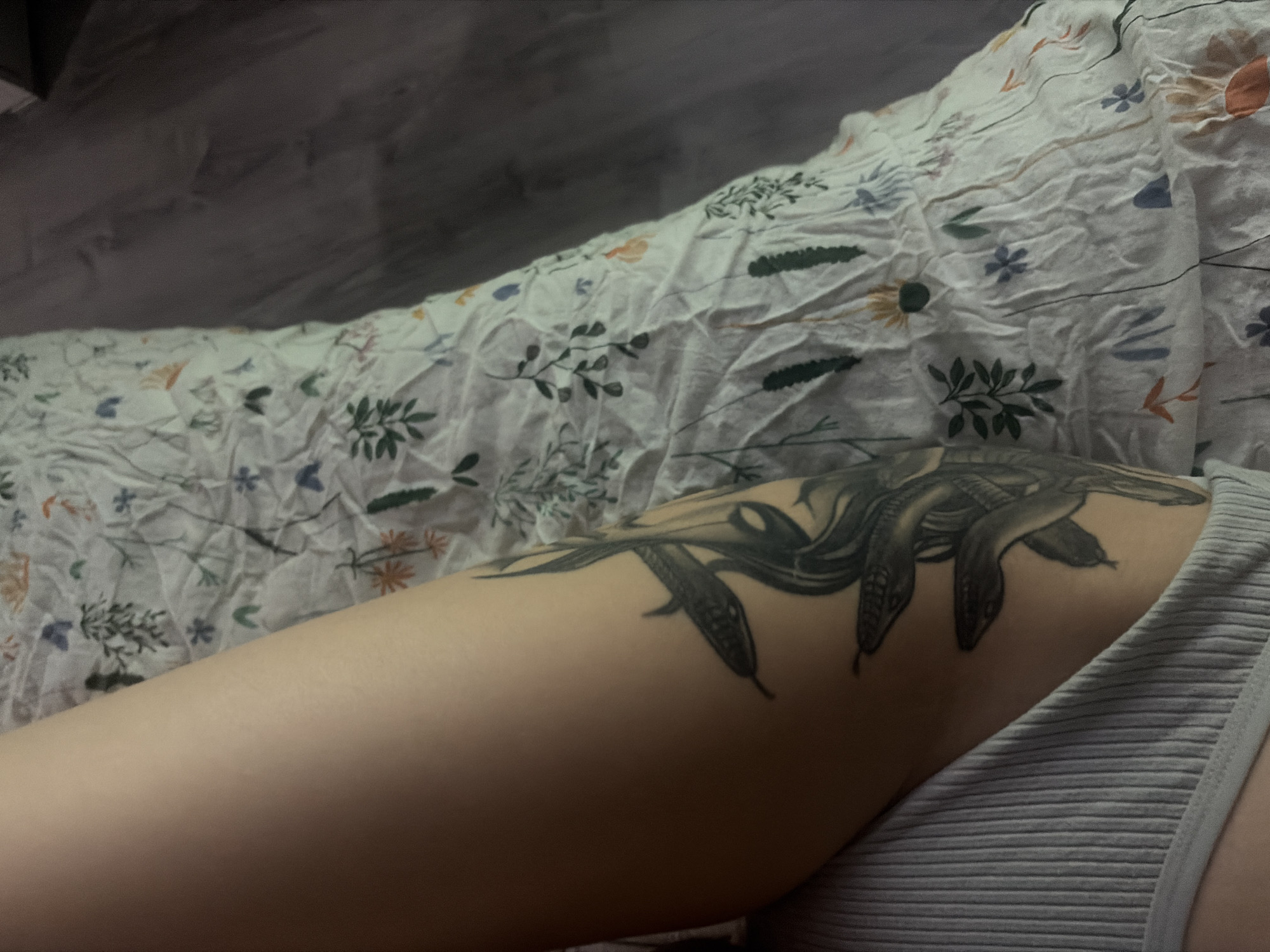 A close-up shot focuses on a person's thigh resting on a patterned bedspread. A snake tattoo is on the thigh. The person is wearing a ribbed tank top. The environment appears to be indoors, with part of a wall visible. There is no indication that the image was taken in Saint Petersburg, Russia.