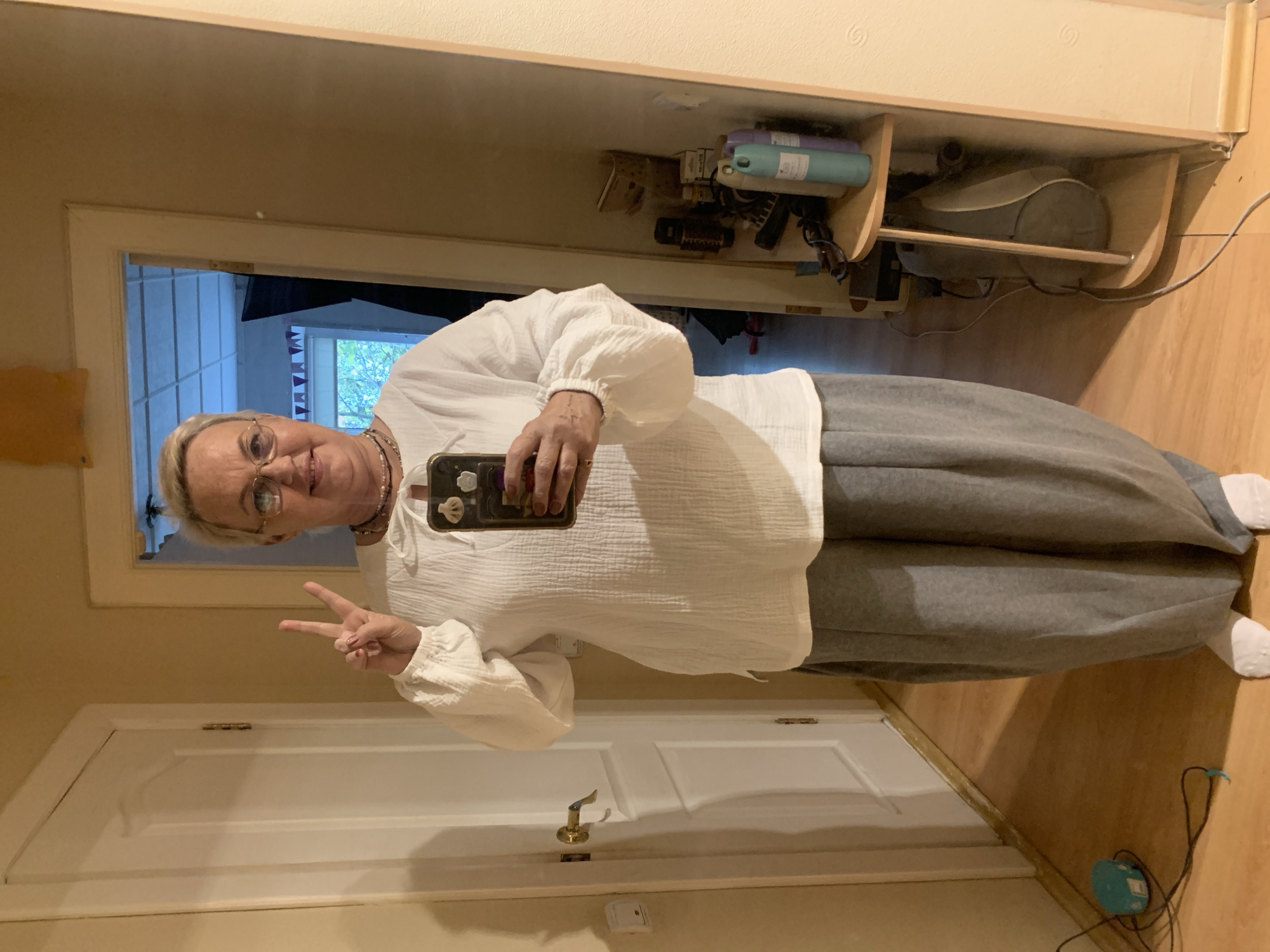 A woman is taking a selfie in a hallway. She is wearing a white top and gray pants and has blonde hair and glasses. She is holding a phone in her right hand and making a peace sign with her left hand. The hallway has a white door and a wooden floor. There is a mirror on the right side of the image, showing shelves with toiletries and a bag. The image appears to have been taken indoors, likely in a residential setting. There is no visible indication that this location is in Chaikovsky, Russia.
