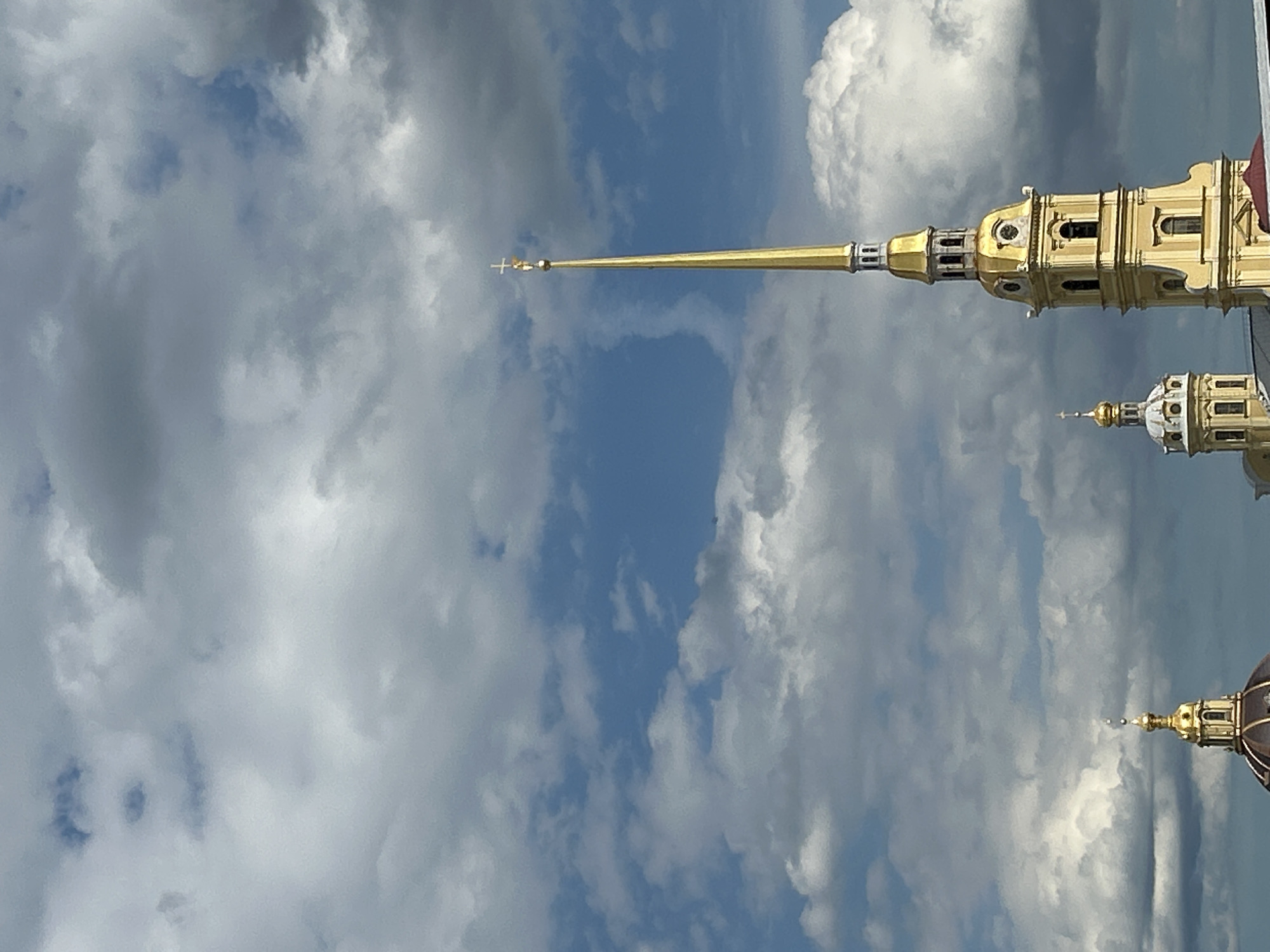 The image features a close-up shot of a golden spire topped with a cross, extending upwards against a backdrop of a cloudy blue sky. The spire appears to be part of a larger architectural structure, with glimpses of ornate golden buildings and domes visible in the background. The clouds are varied, with some appearing dense and gray, while others are bright and white, suggesting a dynamic sky. The sunlight catches the golden elements, making them gleam.
The composition is such that the spire dominates the right side of the frame, with the vast sky occupying the left. The perspective is from below, looking up towards the sky and the structures. The overall impression is one of grandeur and height, likely representing a significant religious or historical building. The weather appears to be a mix of sunshine and clouds, typical of a day with shifting atmospheric conditions. There is no visible text in the image. The scene evokes a sense of awe and possibly a religious or historical significance due to the spire and cross.