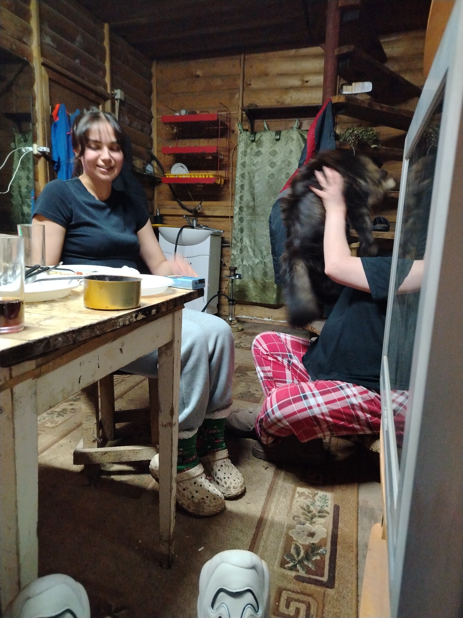 The image appears to be taken indoors, likely in a rustic cabin or cottage-style home with wooden walls and ceilings. The location is Chaikovsky, Russia.
A woman is seated at a weathered wooden table, smiling at something or someone off-camera. The table has dishes and food, including what looks like an open can. She is wearing a dark t-shirt and light-colored sweatpants.
To her right, another person is sitting on the floor, wearing red and white plaid pants and a dark t-shirt. They are holding a medium-sized, dark-colored dog or possibly a small bear.
There is a rug on the floor with floral patterns. A mirror is partially visible on the right side of the frame.
In the background, there's a sink, a red rack, and a curtained window. Stairs can be seen in the upper right corner.