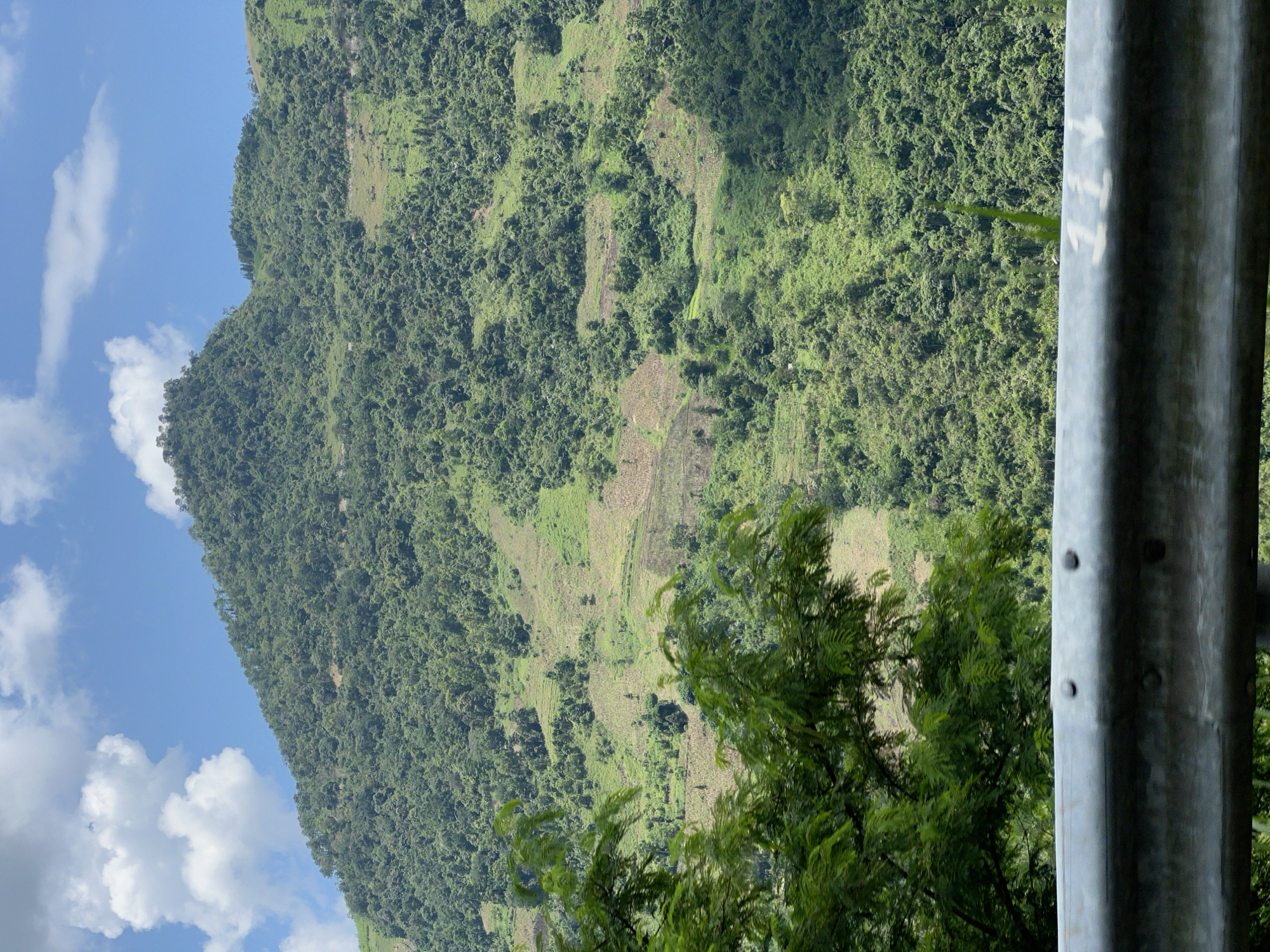 The image shows a lush, green mountain slope under a blue sky with white clouds. The mountain is covered in dense vegetation, with patches of cleared land visible, suggesting agricultural activity or terraced farming. In the foreground, a metal guardrail runs horizontally across the lower portion of the frame, partially obscuring the view. Green foliage from trees hangs over the guardrail, adding depth to the scene. The overall impression is of a rural, mountainous landscape. The location is identified as Minh Tân Commune, Vietnam.