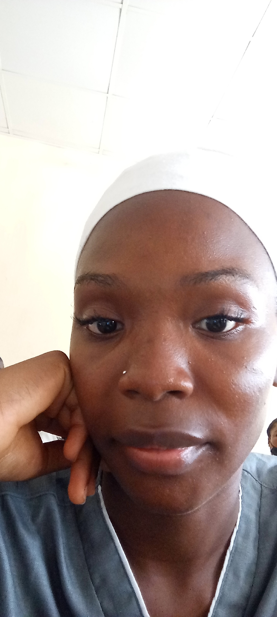 A close-up, slightly angled selfie depicts a dark-skinned woman with a white head covering, potentially indicating a medical or related setting. Her left hand is supporting her face. A small, possibly metallic, nose stud is visible. She's wearing a grey top with a white collar. The background appears to be a plain, light-colored wall or ceiling. There is no location given in the image.