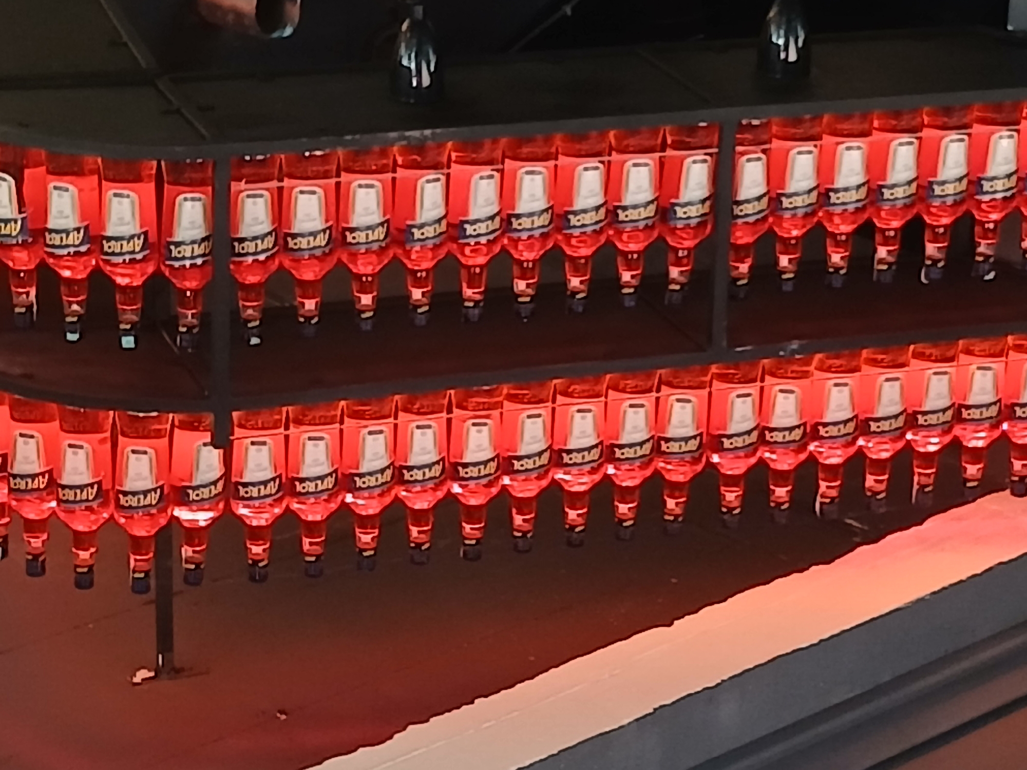 The image shows a shelf stocked with multiple bottles of Aperol. The bottles are arranged in rows on dark shelves, illuminated by red lighting from above. Each bottle is clear glass, filled with orange liquid, and has a white label with the brand name "APEROL" clearly visible. The labels also feature a black band and a white rectangular section. The bottles have black caps and a thin black neck. The background shows a dark, possibly wooden, ceiling with some structural elements. There are no people or other discernible objects in the frame, other than the bottles themselves. The scene appears to be in a bar or retail establishment, given the organized display of alcoholic beverages. The location is unknown.