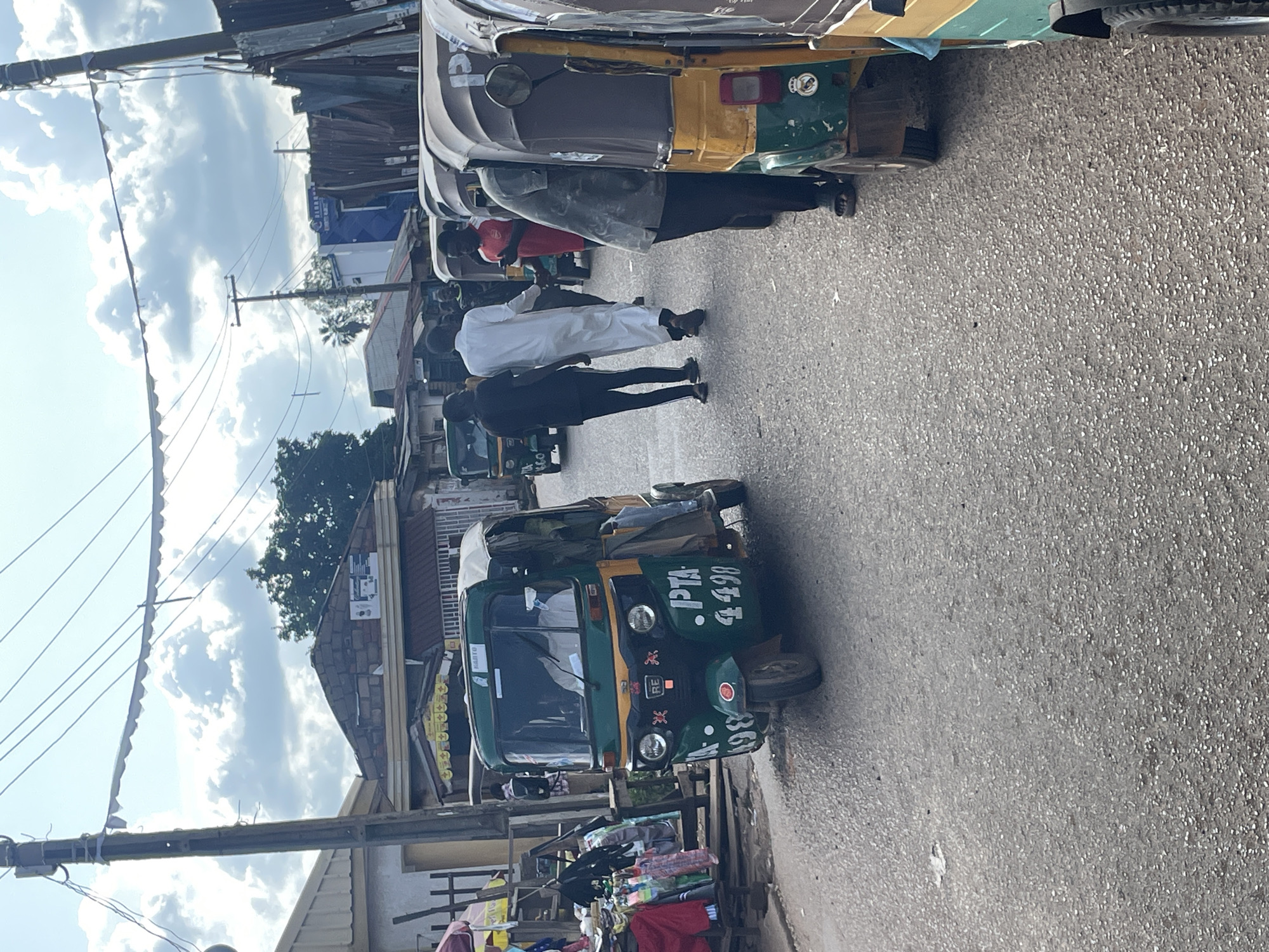 In Gura Top, Nigeria, a street scene unfolds with several auto-rickshaws, commonly known as "keke napeps," lining a paved road. The foreground is dominated by a green and yellow keke napep with the markings "PTA 4498" on its side, a driver visible inside. To the right, a line of similar vehicles extends into the background. A few individuals are present; two men in dark clothing are walking in the street, and another man in a white robe is also walking. The environment appears to be a busy urban area with buildings and overhead power lines visible. The sky is partly cloudy.