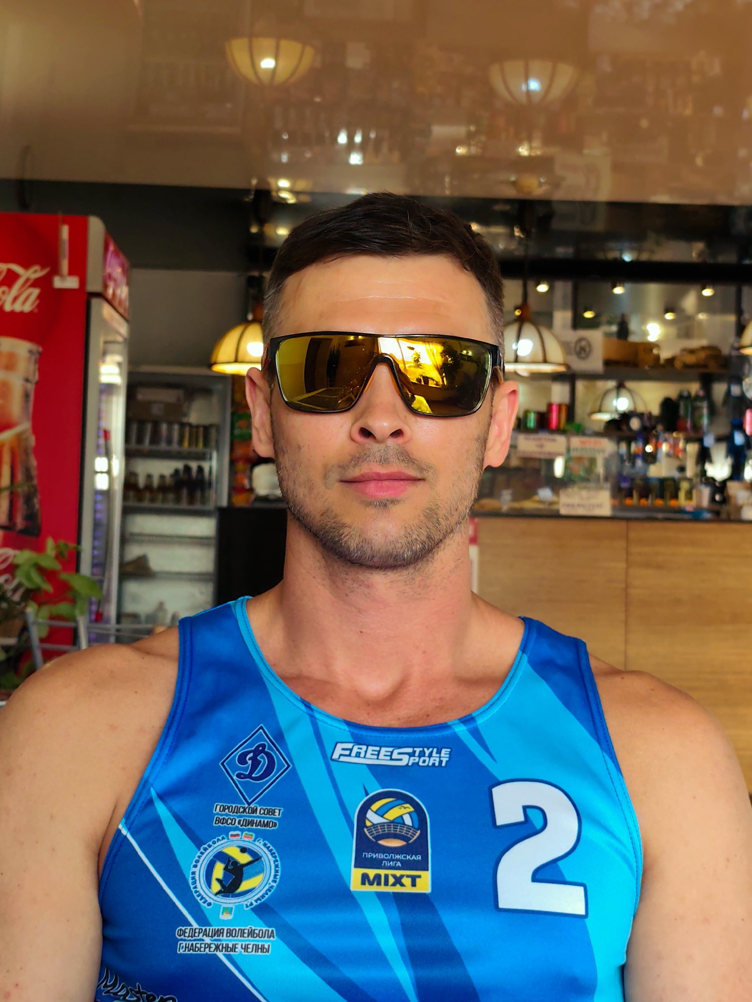 A medium shot captures a man wearing mirrored sunglasses and a blue athletic tank top, likely taken in an indoor setting in the city of Нижний Пальник, Russia. The man has short, dark hair and a stubble beard. His tank top features various logos and text in Russian, including "Г.НАБЕРЕЖНЫЕ ЧЕЛНЫ" (Naberezhnye Chelny city), suggesting a sports team or event affiliation. The number "2" is prominently displayed on the right side of his tank top. The reflection in his sunglasses reveals a glimpse of the surrounding environment, including light fixtures and potentially other people. In the background, a red Coca-Cola refrigerator stands next to a counter displaying various objects, hinting at a café or restaurant setting.