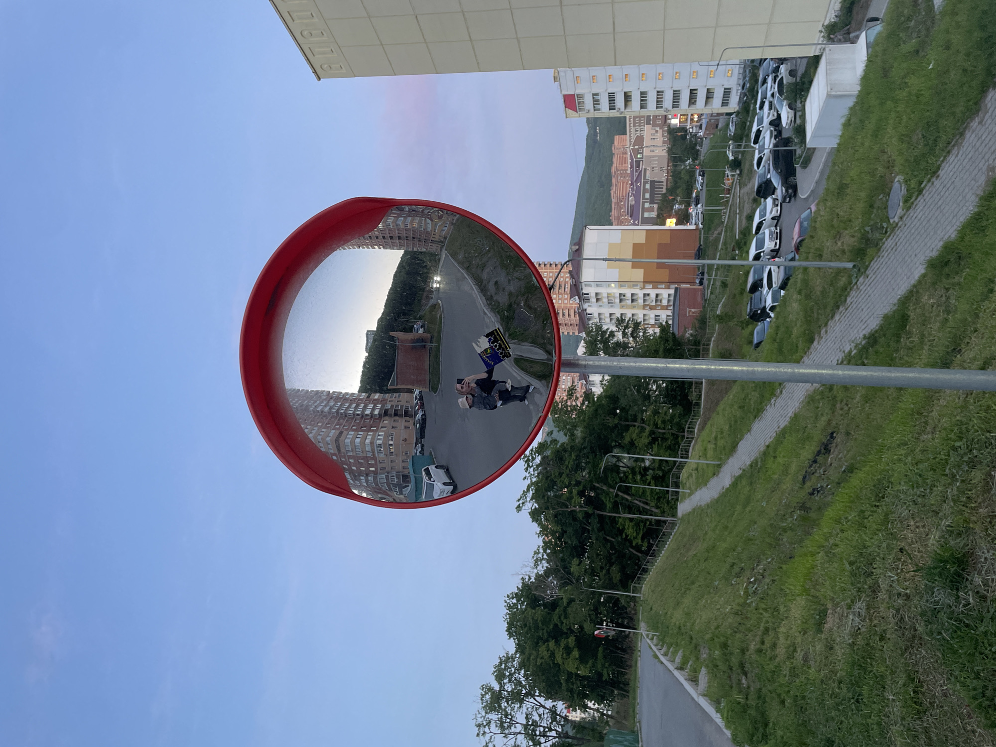 A convex mirror reflects a scene in Vladivostok, Russia. The reflection shows two individuals, presumably taking a selfie. Surrounding the mirror in the actual scene are buildings, greenery, and a road. A parking area with numerous cars is also visible. The sky appears to be transitioning from day to dusk.
