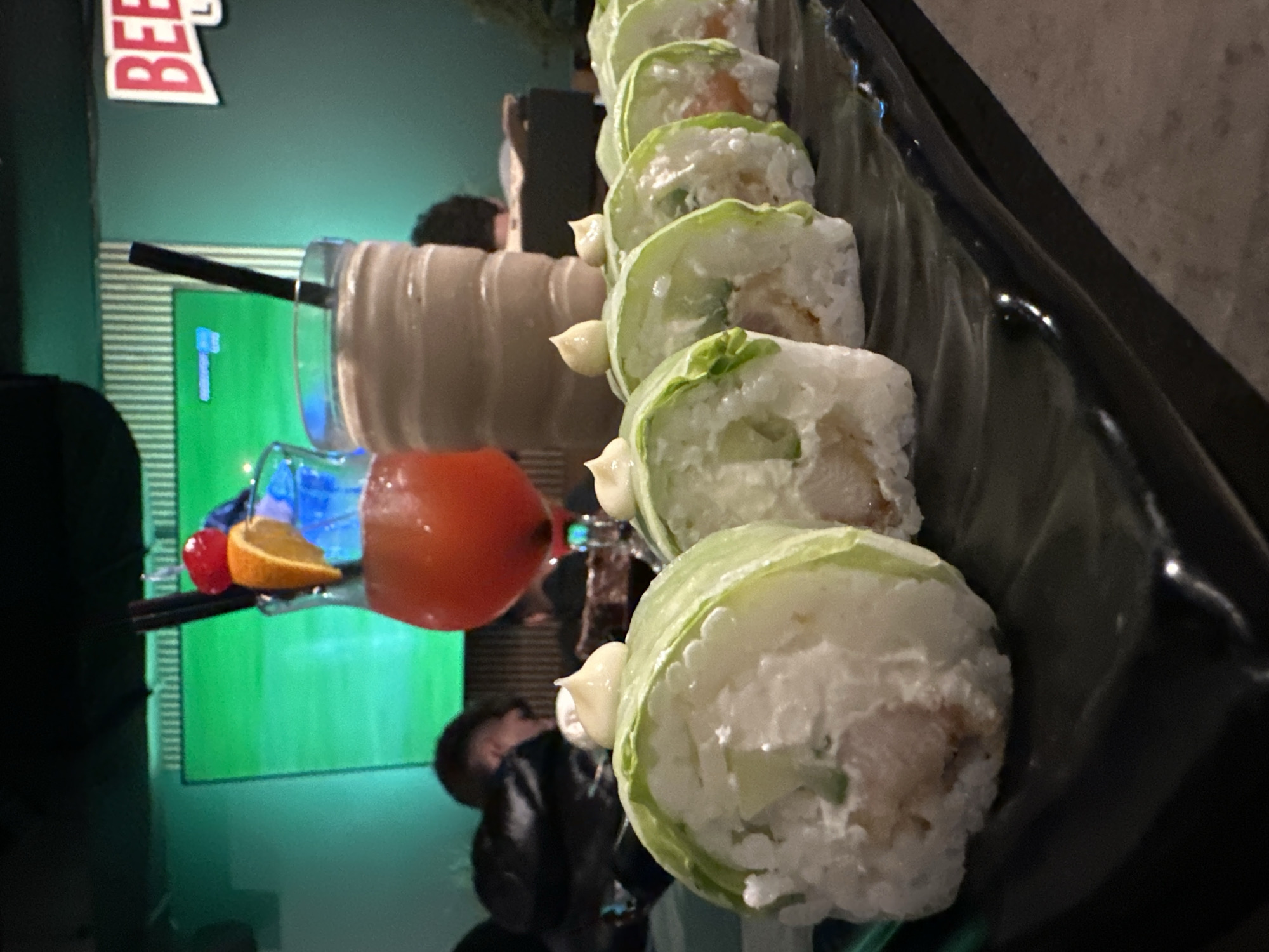 The image shows a plate of sushi rolls, with white rice and light green wrapping, arranged on a black dish. A glass of dark beige liquid with a black straw stands behind the sushi. A glass of red liquid garnished with an orange slice and cherry stands beside it. A green wall with white stripes is visible behind the glasses. A person is seated behind the drinks, with a partial view of their head and shoulders. Another person is blurred in the lower left. The image was taken in Burgas, Bulgaria.