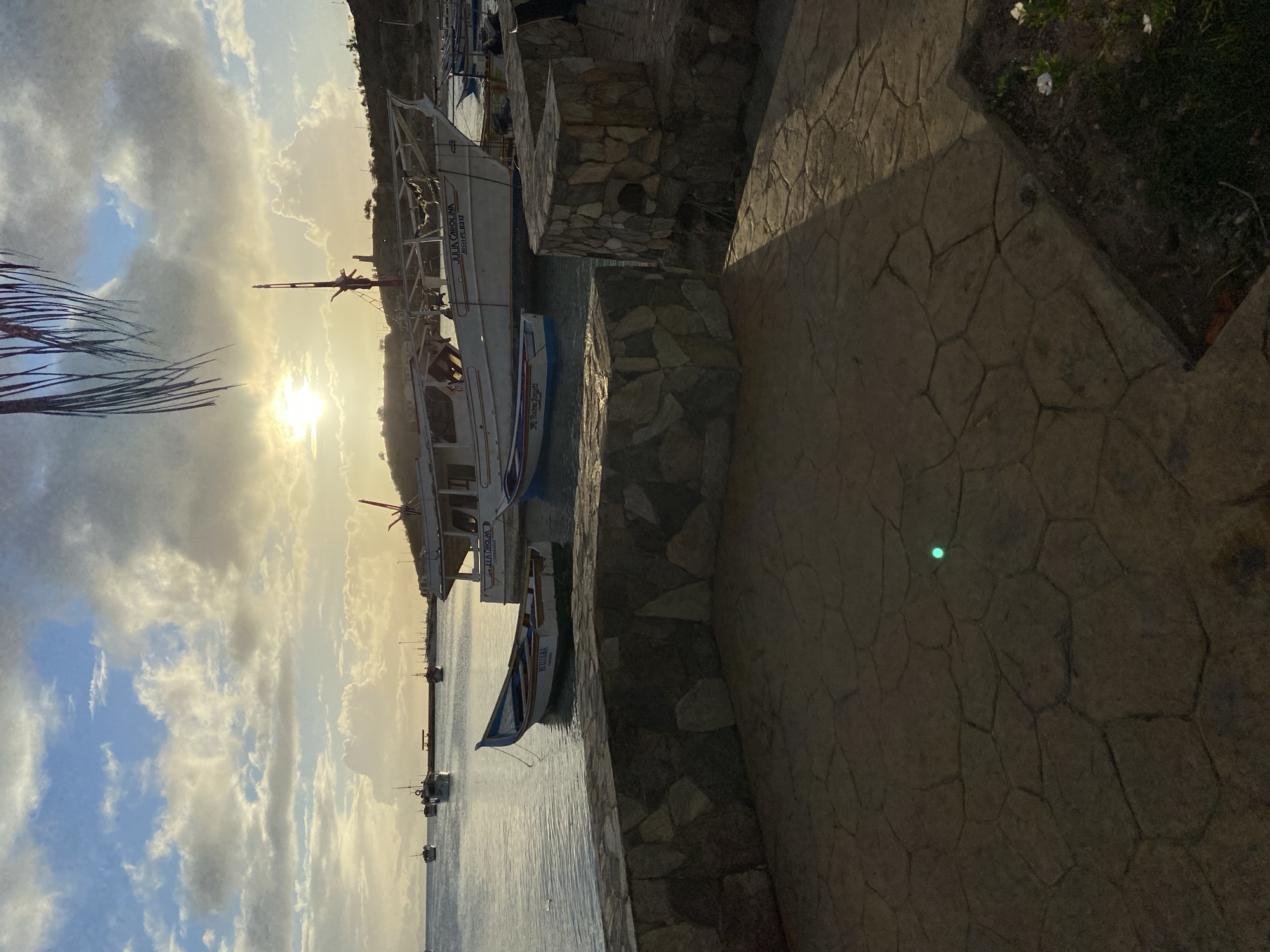 The image depicts a tranquil harbor scene in Juan Griego, Venezuela, during what appears to be sunset or sunrise. In the foreground, a stone-paved patio area casts shadows from a nearby structure, with a single bright green light visible on the ground. A low stone wall separates the patio from the water, where several boats are moored.
Dominating the midground are two boats. A larger white boat, named "JULIA CAROLINA" with registration "ARGH-PE 0312," is prominently displayed. Beside it, a smaller, narrower boat with blue trim is tethered. In the background, more boats are scattered across the calm, reflective water, with a distant harbor or pier visible. The sky above is a mix of blue and dramatic clouds, with the sun's rays breaking through, creating a warm, hazy glow. The shoreline behind the harbor features a hilly, sparsely vegetated landscape. A few palm fronds hang from the top right of the frame, adding a tropical element to the scene. No people are visible in the image.