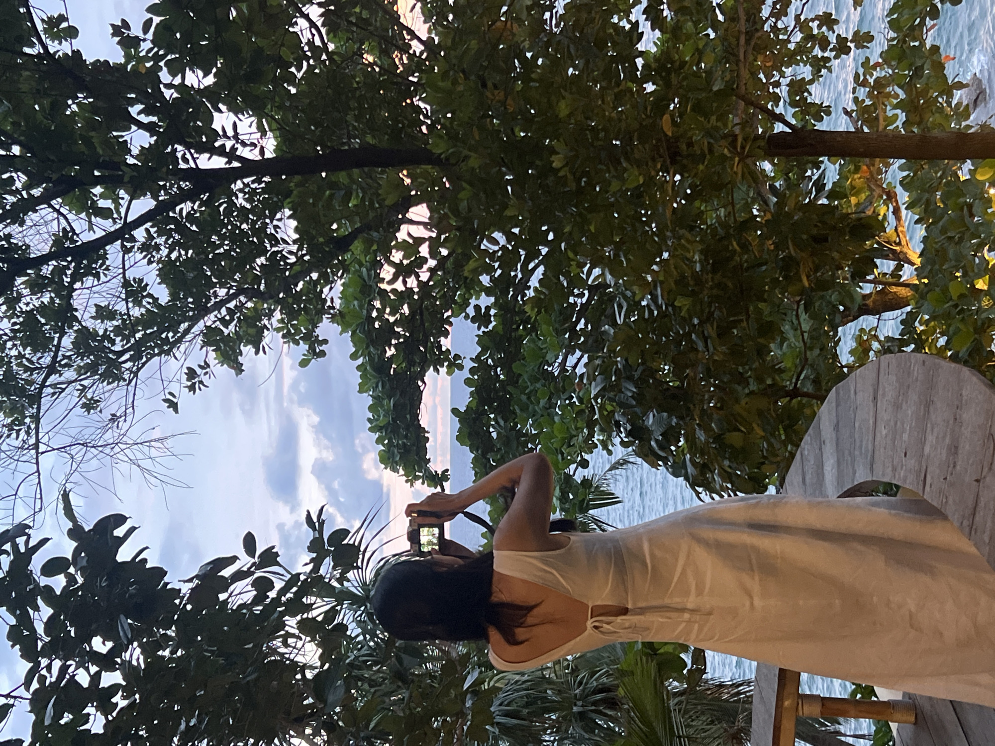 A person, seen from the back, wearing a white dress, is standing and holding a camera up to their face, appearing to take a photograph. They have long, dark hair and are positioned on a wooden platform or table. The setting is outdoors, with lush green foliage from trees and plants surrounding them. Beyond the greenery, a body of water, likely the ocean, is visible, with gentle waves and a hint of the sky above. The sky is a mix of blue with scattered clouds, and the lighting suggests it might be late afternoon or early evening, with a soft, diffused light. The overall atmosphere appears serene and tranquil. The location is Karon, Thailand, which aligns with the tropical vegetation and ocean view.