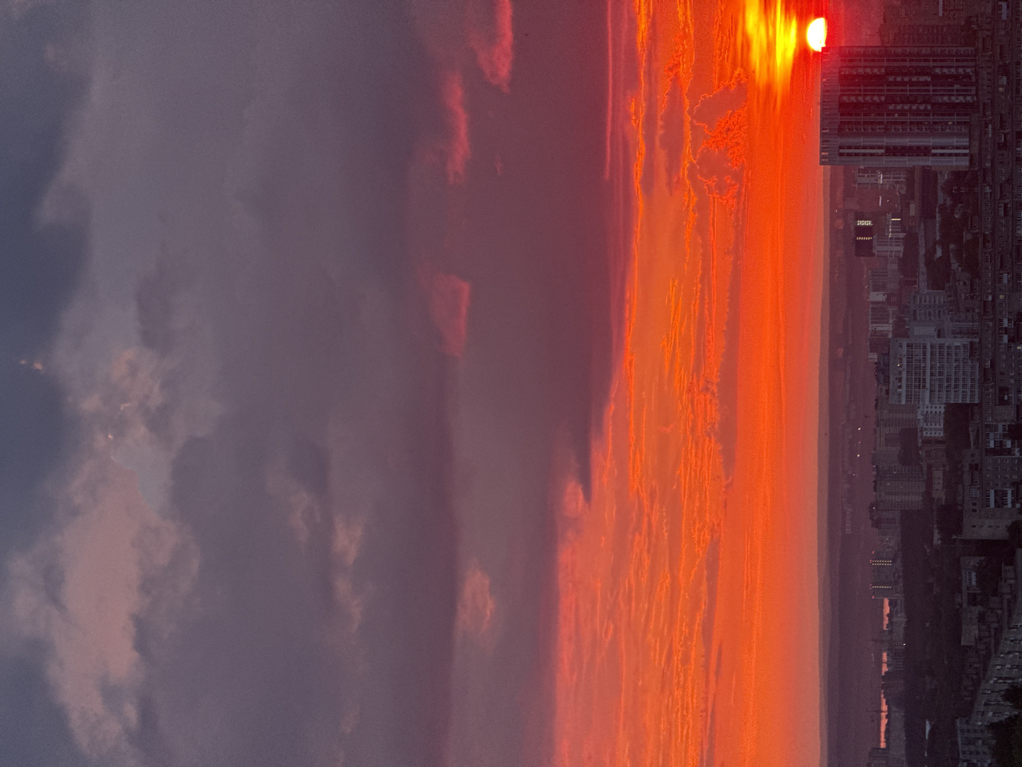 The image captures a dramatic sunset over the city of Savenki, Russia. The sky is a vibrant canvas of fiery orange and deep purple, with clouds tinged by the setting sun's intense hues. Below the spectacle of the sky, the urban landscape unfolds. Buildings of varying heights, predominantly residential blocks, stretch across the horizon. A large, modern building with a distinctive vertical pattern stands out among the others, its façade reflecting the last rays of the sun. The lower parts of the city are cast in shadow, creating a stark contrast between the illuminated sky and the darkening city. The scene is devoid of any visible people or specific actions, presenting a serene, yet powerful, depiction of an urban environment at dusk.