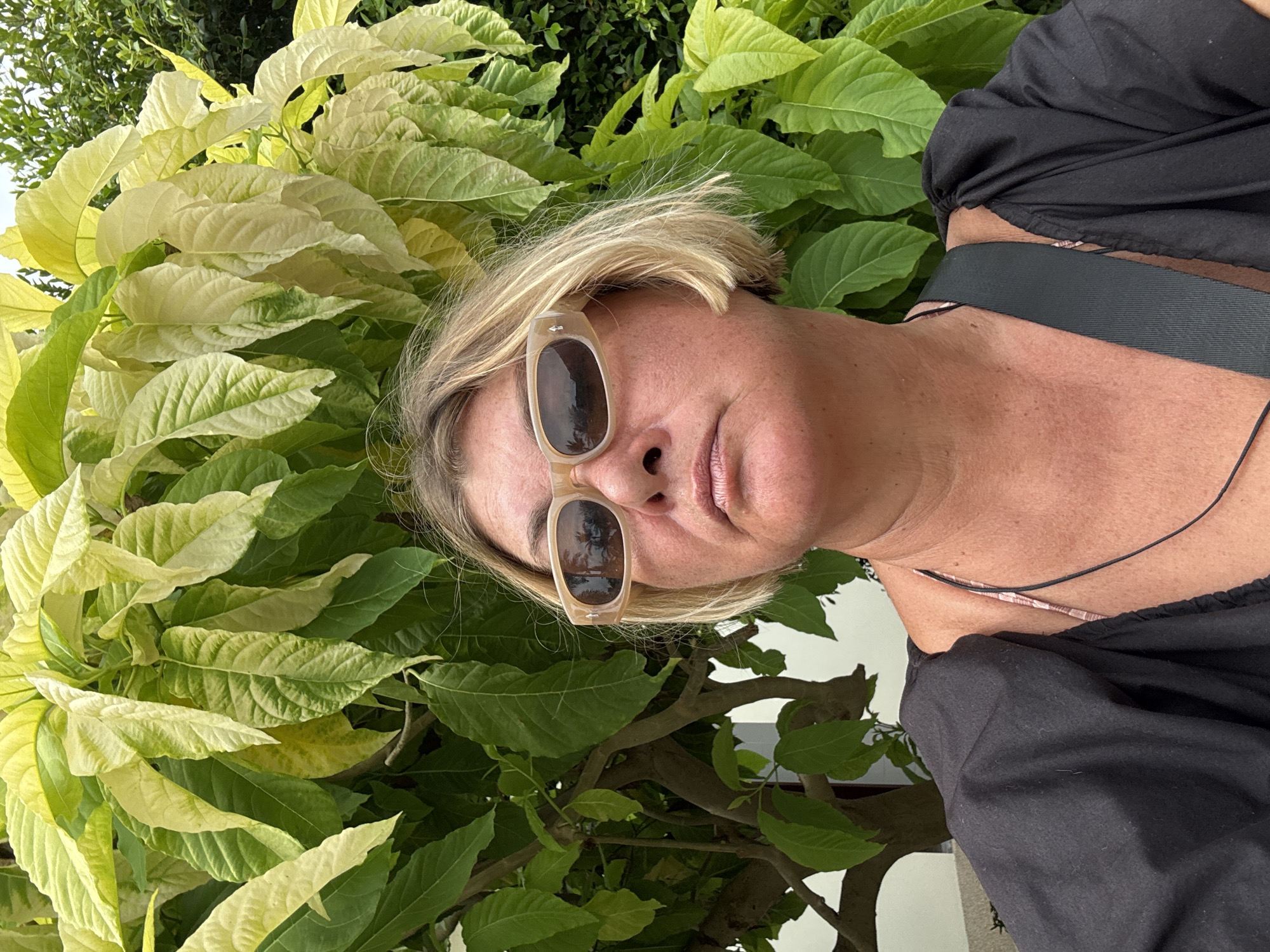 A woman with short blonde hair and sunglasses is framed by lush green foliage with some variegated leaves. She is wearing a black top. The photo appears to be taken outdoors, potentially in Baan Tai, Thailand, given the prompt's suggestion.