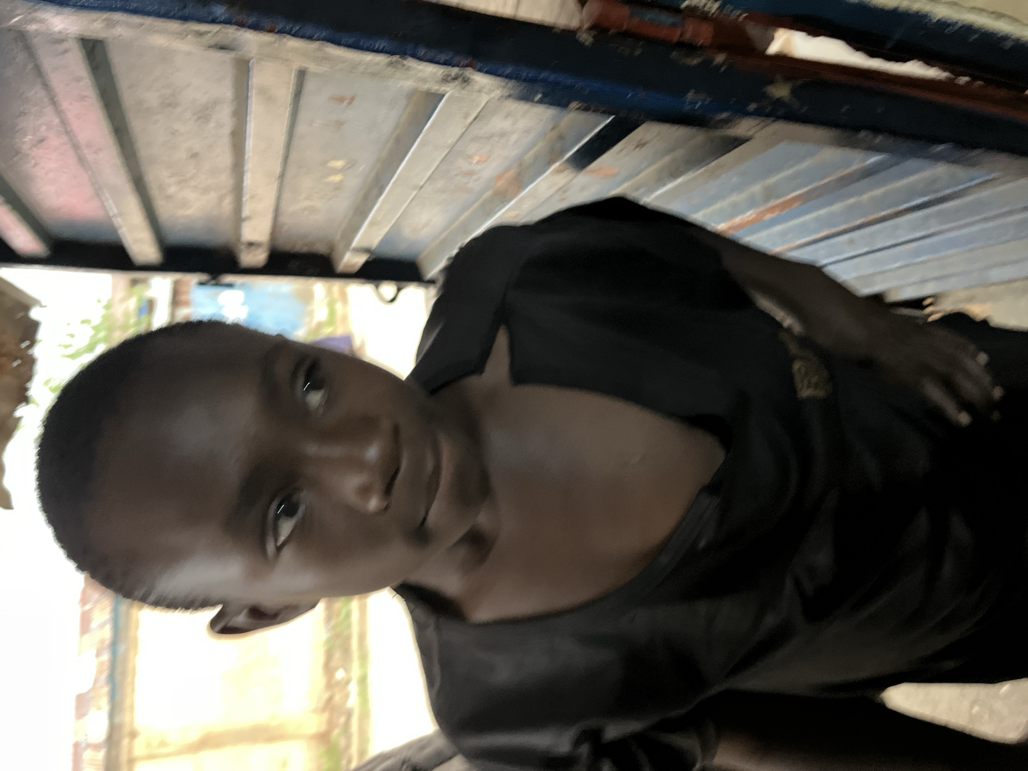 The image presents a close-up view of a young person with dark skin and short hair, wearing a dark-colored shirt. The person appears to be looking towards the camera with a neutral expression. The background shows glimpses of an exterior environment, possibly a street scene with buildings and foliage, but it is out of focus. Overhead, there is a structure consisting of parallel metal bars, possibly part of a vehicle or a building frame. The scene does not provide enough specific details to definitively confirm the location as Jos, Nigeria.