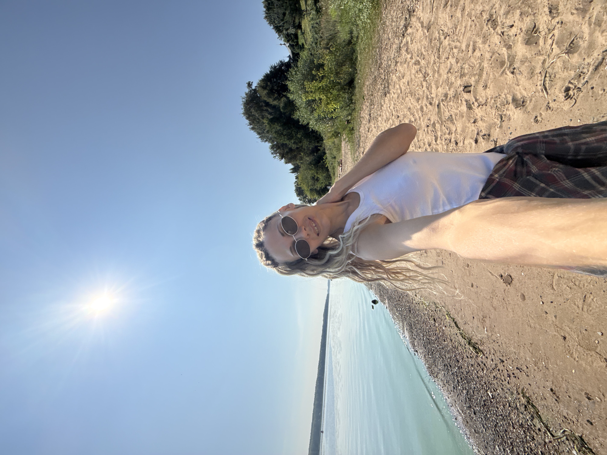 A woman with blonde, braided hair and sunglasses takes a selfie on a sandy beach. She is wearing a white tank top and a plaid shirt tied around her waist. The sun shines brightly in a clear blue sky. To her left is a body of water with a rocky shore, and to her right are trees and green foliage. The location appears to be the city of Rusanovo, Russia.