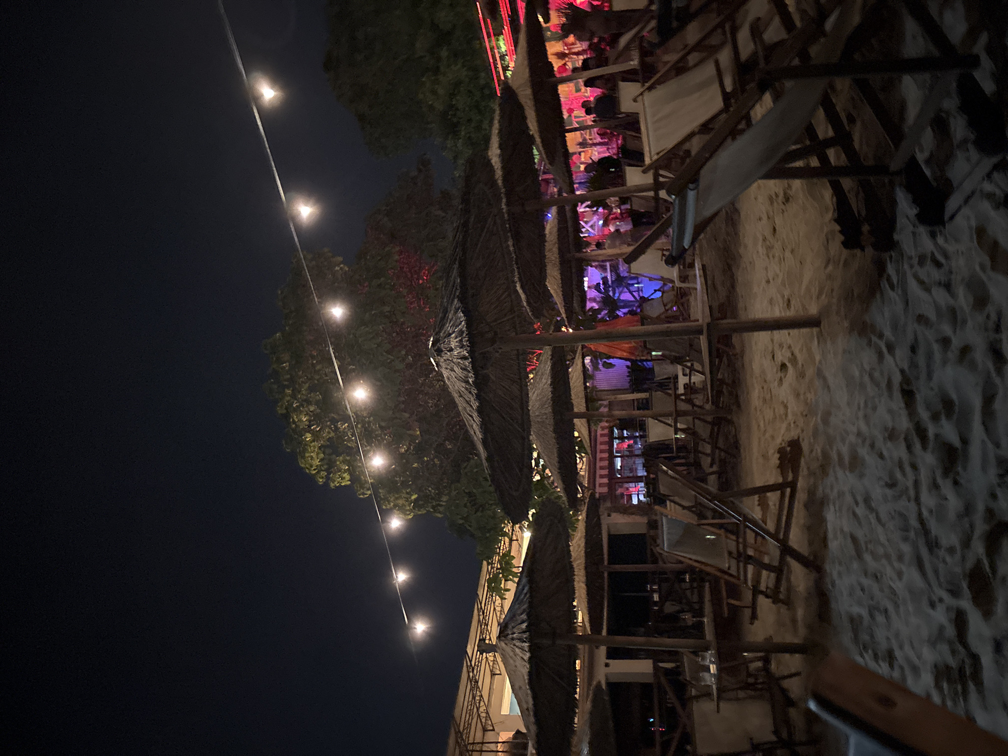 The image displays a nighttime scene at an outdoor establishment situated on a sandy ground, characteristic of a beach club or open-air restaurant in Varna, Bulgaria, likely along its coastline such as the South Beach or within the Sea Garden area. The sky is dark.
In the foreground and mid-ground, numerous straw-thatched umbrellas are erected across the sandy expanse, casting shadows. Multiple white canvas beach chairs, some folded and some open, are arranged around small wooden tables. On one table in the left mid-ground, two glasses are visible.
A string of incandescent bulbs hangs diagonally across the upper portion of the frame, providing overhead illumination to the area. Dark foliage from trees is visible in the background, some appearing to be partially illuminated by ambient light.
In the mid-ground and background, several individuals are congregated, particularly in areas illuminated by vibrant artificial lighting, including red, purple, and pink hues emanating from unseen structures. These lights suggest a lively, social atmosphere. In the left background, a multi-story building with an open metal roof structure is partially visible. Further to the right in the background, other structures with intense colored lighting indicate a vibrant gathering space. Individuals appear to be standing and socializing in these brightly lit zones.