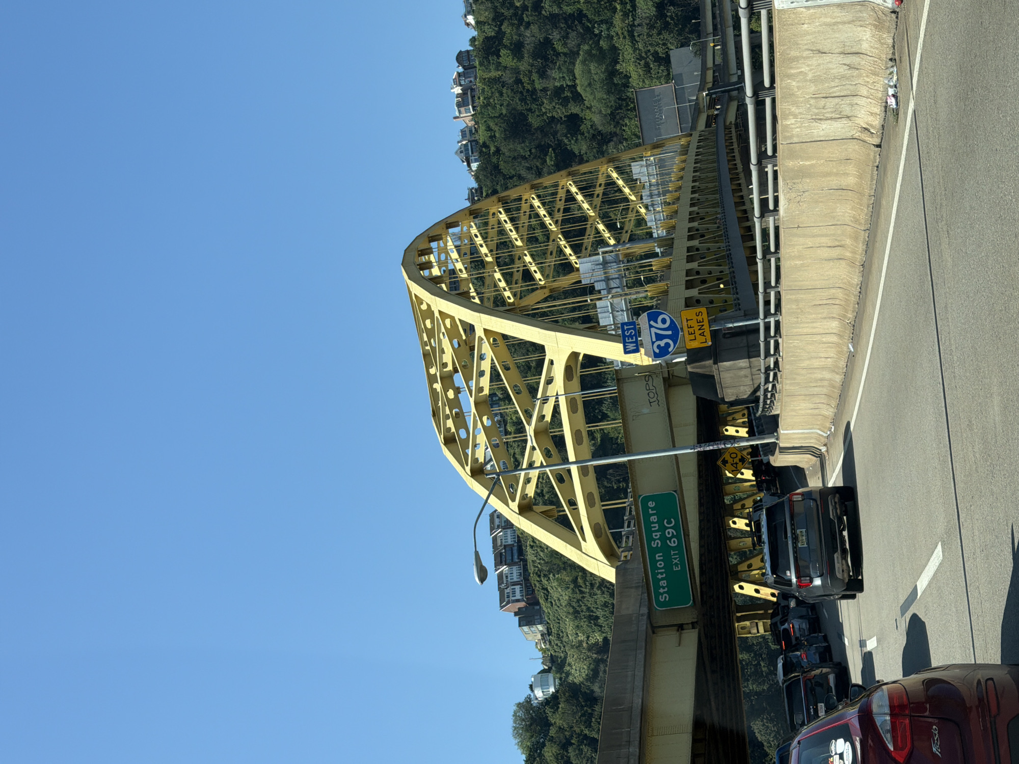 The image depicts a traffic scene on an elevated highway in Pittsburgh, Pennsylvania, under a clear blue sky. A large yellow steel arch bridge dominates the upper portion of the frame. Below the bridge, a green highway sign indicates "Station Square EXIT 69C". To the right of the bridge's arch, a blue circular sign with "WEST 376" and a yellow rectangular sign with "LEFT LANES" are visible.
On the highway, a line of vehicles is moving slowly. In the foreground, a red car is visible from the rear. Behind it, a gray SUV is in the center lane, followed by several other cars in adjacent lanes. The highway is flanked by a concrete barrier. Beyond the barrier and the highway, lush green trees cover the hillside, and houses are perched on top of the hill. The overall impression is of a busy commute during daytime.