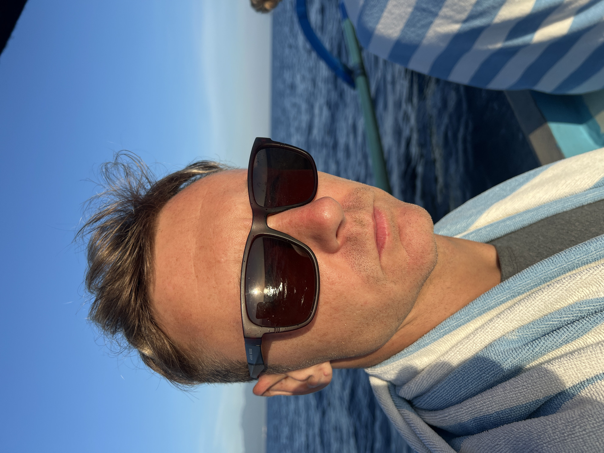 A fair-skinned man wearing sunglasses and draped with a blue and white striped towel is in a boat on the water. The sky is visible in the background. The location is specified as Seririt, Indonesia, though there is no evidence in the image to support this.