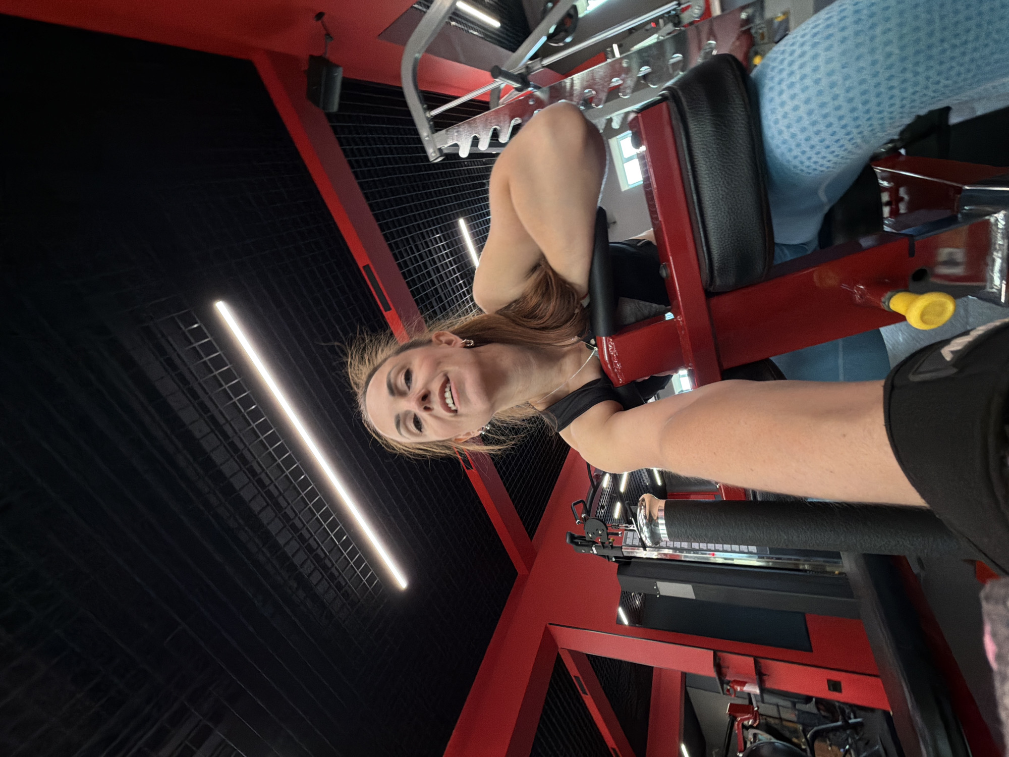 In a gym located in Birobidzhan, Russia, a woman with brown hair and light skin is captured from a low-angle perspective. She is wearing a black tank top and light blue patterned leggings. The woman is seated on a red gym machine with black padding, her arms are resting on the padding, and she is looking towards the camera with a slight smile. The environment is characterized by a black ceiling with grid panels and recessed lighting, and red gym equipment and structures. A portion of a person's arm, wearing black fitness gloves and a black sleeve, is visible in the foreground, suggesting the photo was taken by the person.