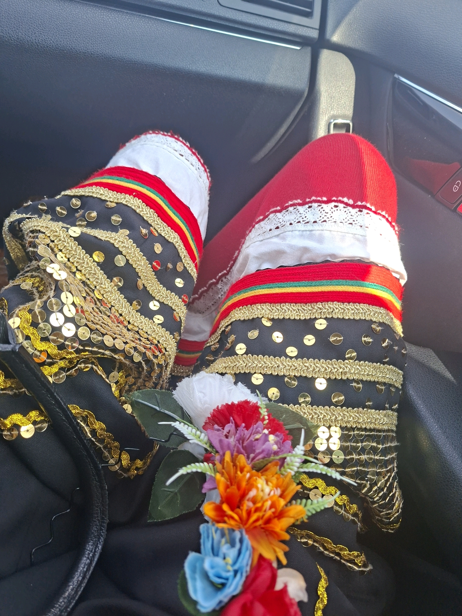 A close-up shot captures the lower legs of a person wearing traditional, heavily ornamented attire. The garments are black, adorned with gold trim, sequin embellishments, and colorful stripes featuring red, yellow, and green. The upper portion of the legs is covered by white fabric with white lace trim, and then a solid red fabric. A small artificial floral arrangement with red, orange, purple, and blue flowers sits on the black fabric near the person's left ankle. The background suggests the interior of a vehicle, with dark textured upholstery visible, including a car door handle and a portion of the dashboard. The lighting indicates it is daytime.