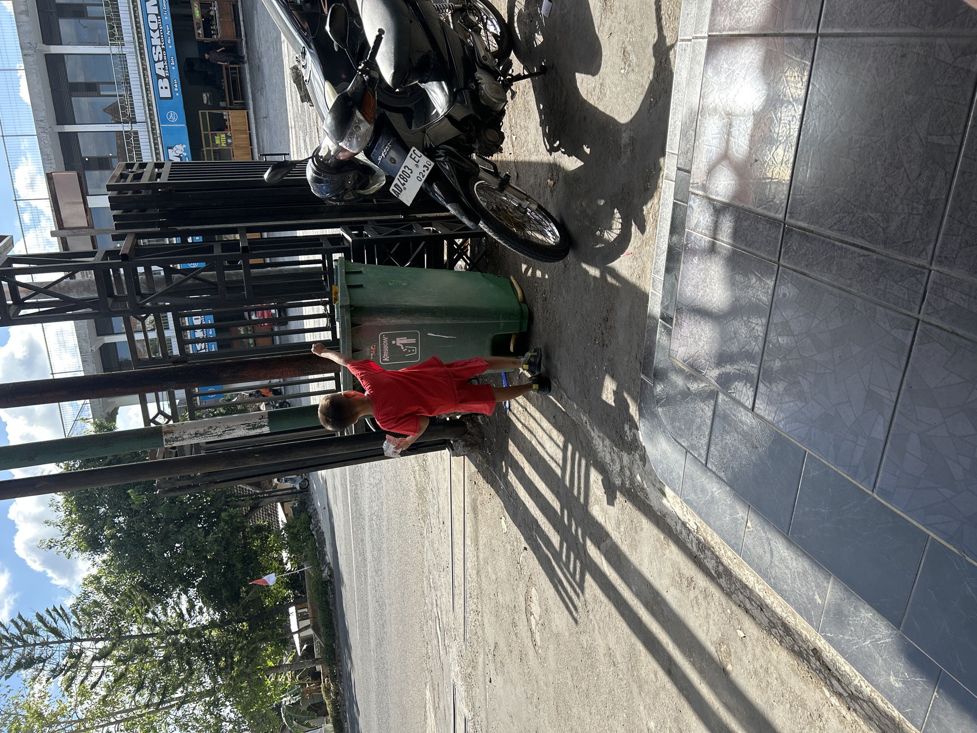 A young boy in a red shirt and shorts stands next to a green trash can, holding a small object in his hand. He appears to be interacting with the trash can, possibly about to discard the object. The boy is positioned on a sidewalk made of gray tiles with a patterned design, casting long shadows due to the bright sunlight. Behind him, a black motorcycle is parked. In the background, there is a street, trees, and buildings. One building features a sign that says "BASKOM" and another smaller sign with a blue circular logo. The sky above is clear with some white clouds. The overall scene suggests an outdoor urban environment, likely in a city, given the presence of buildings, a street, and a parked motorcycle. The image is taken in Wonosari, Indonesia.