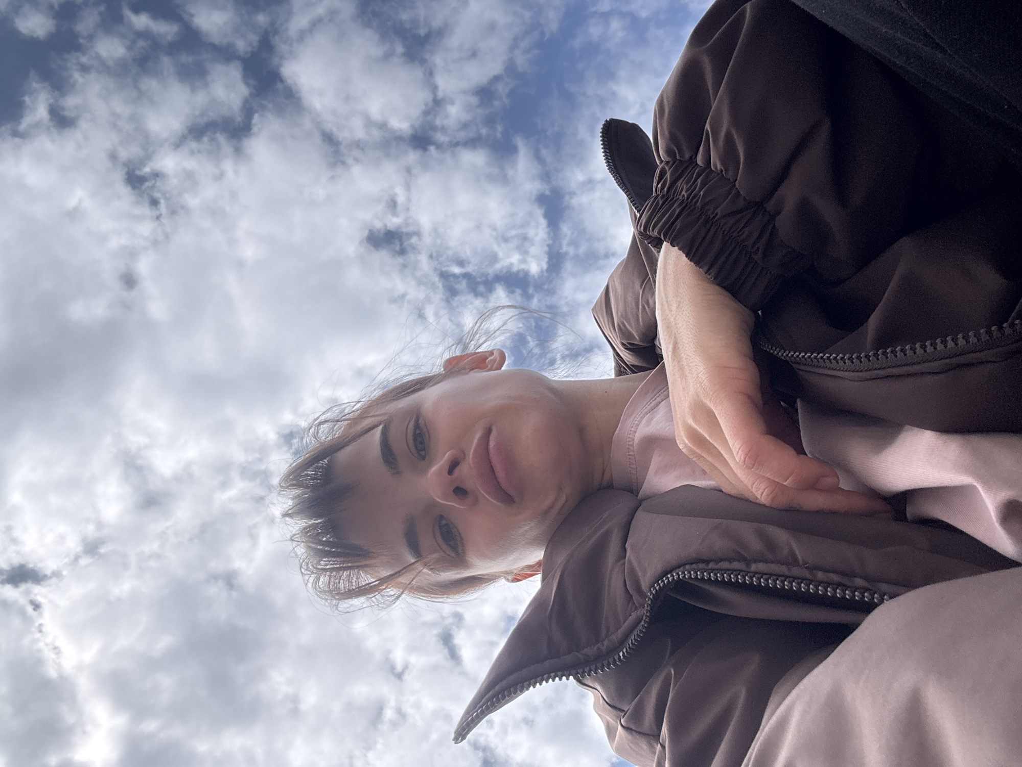 The image is a low-angle, rotated portrait of a young woman, taken from below, looking upwards towards the sky. The woman's face is positioned in the left-center, tilted towards the top-right, as she looks directly down into the camera. She has light-colored hair, slightly tousled, and her expression is neutral to subtly contemplative, with her lips slightly parted.
She is wearing a light pink or peach-colored top under a dark brown or taupe puffer jacket, the texture of which is visible, along with a prominent zipper. Her right hand is visible, resting gently on her jacket, suggesting a relaxed posture.
The background is a bright, partly cloudy sky, featuring patches of clear blue interspersed with numerous soft white and grey clouds. This indicates an outdoor setting during daytime with fair weather. The overall composition suggests a self-portrait or a close-up taken from a very low vantage point. No text is visible in the image. The scene is set in what is understood to be Rostov-on-Don, Russia.