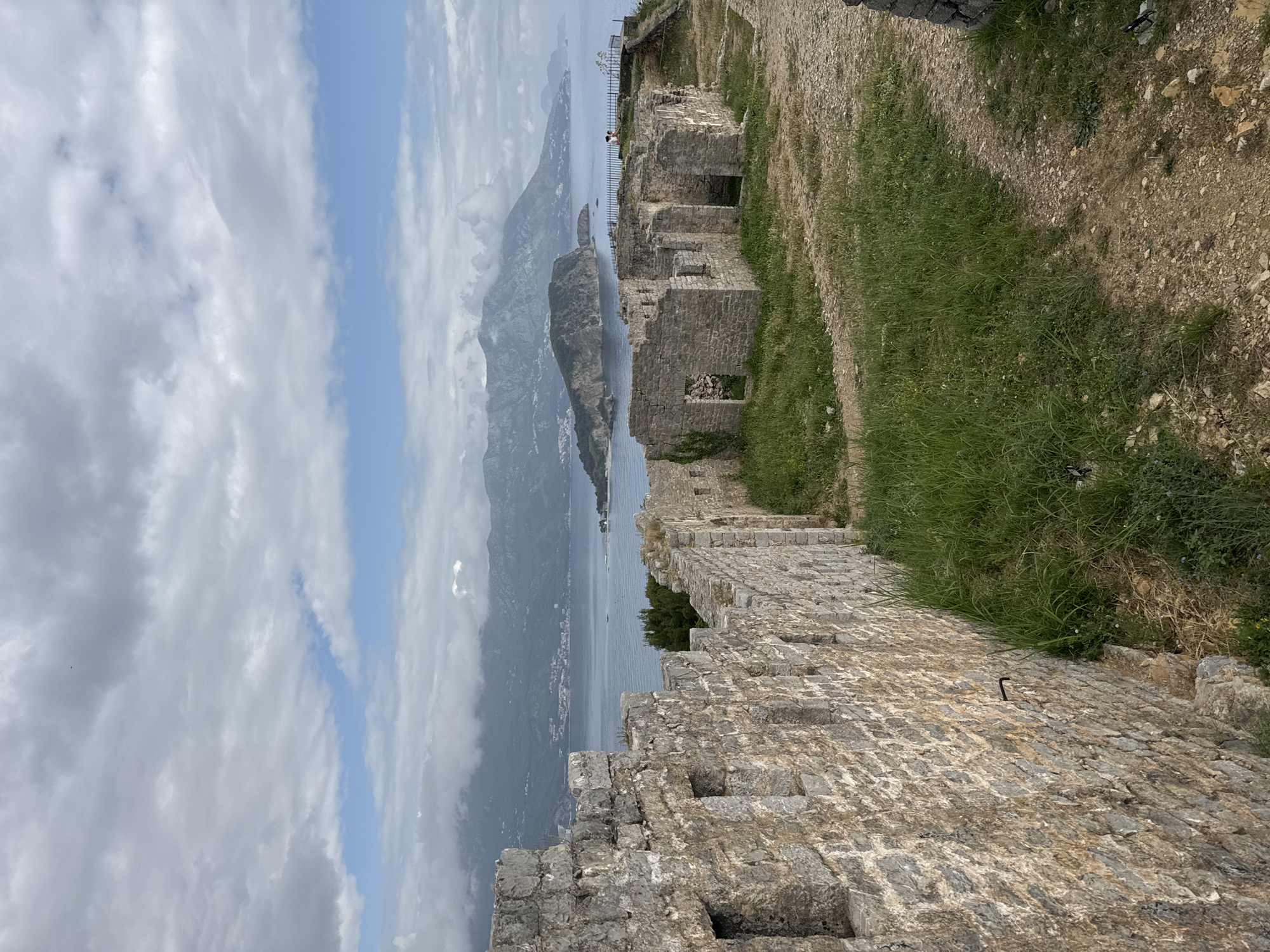 The image shows a view from the Citadel in Budva, Montenegro. The foreground consists of a stone wall and the ruins of buildings, with grass growing between the stones. Beyond the Citadel, there is a view of the Adriatic Sea, a mountainous island, and the coastline, which fades into the horizon under a sky filled with clouds.