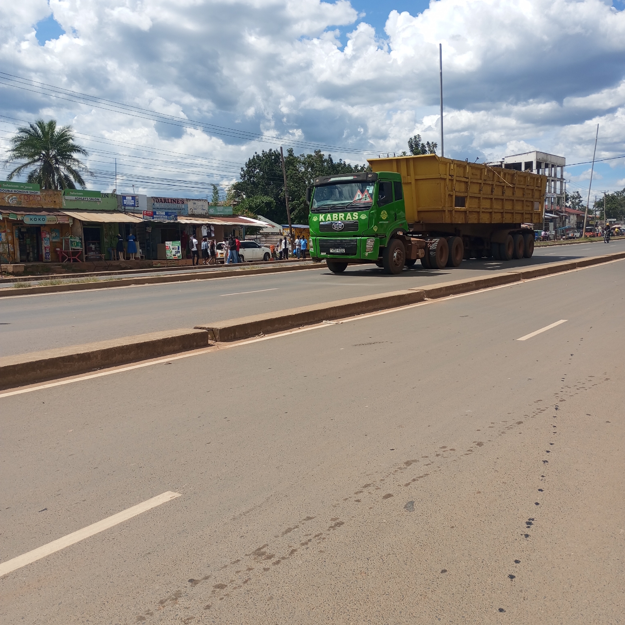 The image shows a green and yellow Kabras truck driving on a paved road in Sheywe ward, Kenya. The road has a center divider and white lane markings. On the left side of the road, there are several small shops including "KOKO", "ASHLEY'S SALON", and "DARLINE'S". People are standing and walking near these shops. The sky is partly cloudy with a few scattered buildings visible in the background. There are dark spots on the road surface in the foreground.
