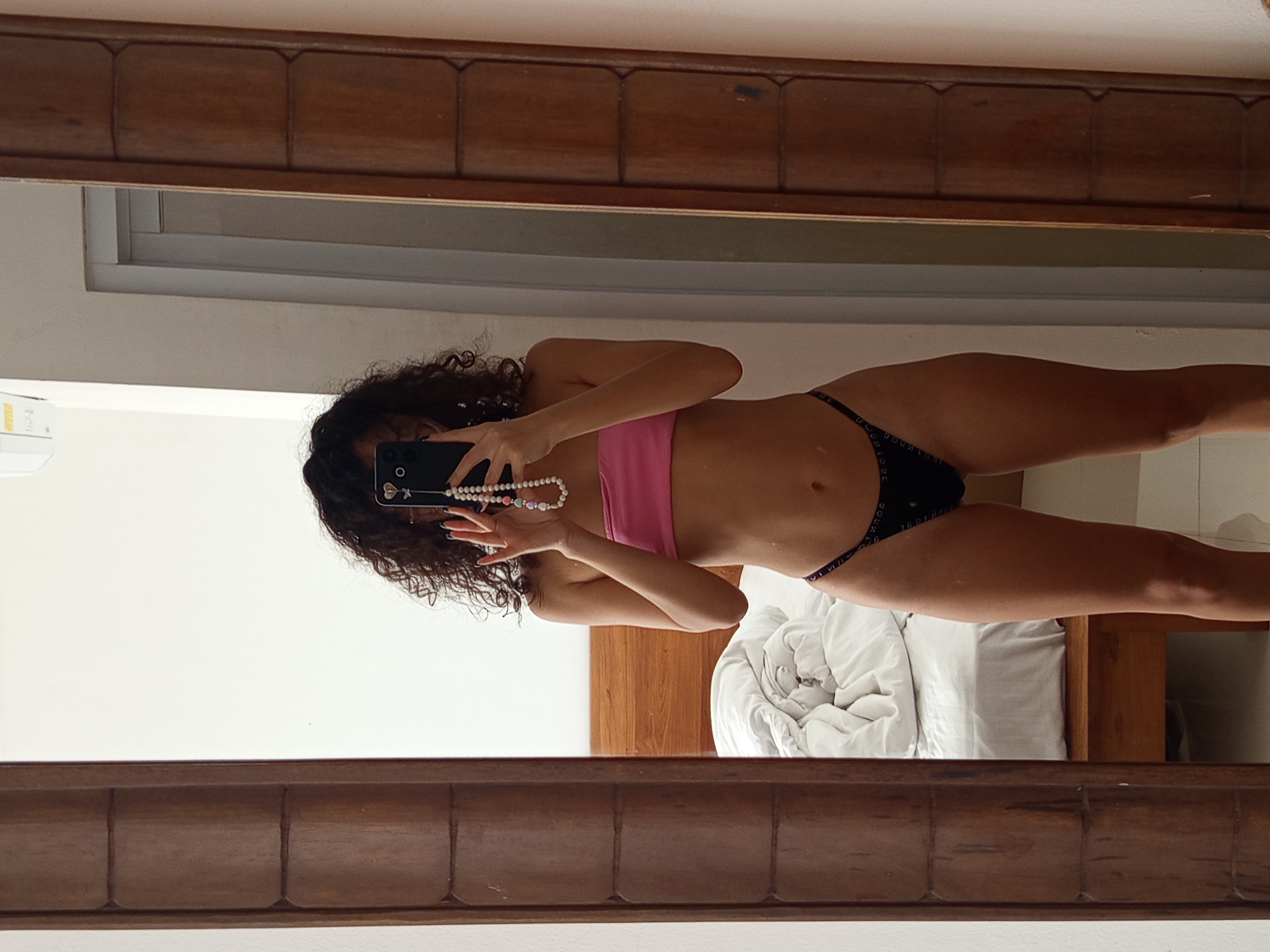 The image is a mirror selfie of a woman in a pink bandeau top and black bikini bottoms. She is holding a black smartphone in front of her face, obscuring her features. The mirror is framed with dark wood. The reflection shows a bed with white bedding and a wooden headboard. The room appears to be a hotel room, potentially located in North Kuta, Indonesia. The wall behind the woman is white.