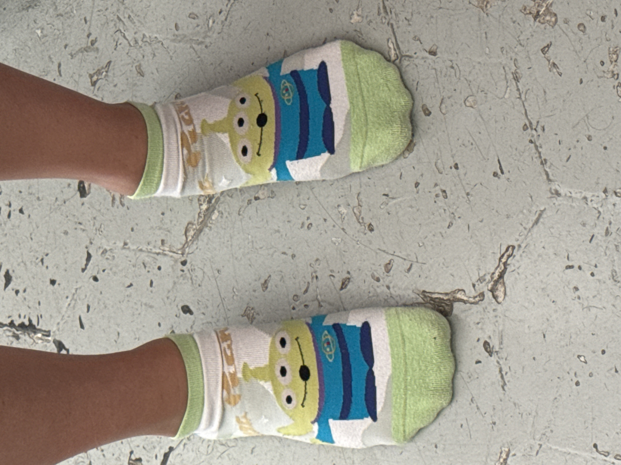 The image shows a pair of feet wearing white socks with green toes and cuffs. The socks feature a design of a three-eyed alien character. The feet are resting on a light gray concrete surface with irregular dark markings. There is no further evidence to suggest the location is in Puebla, Mexico.