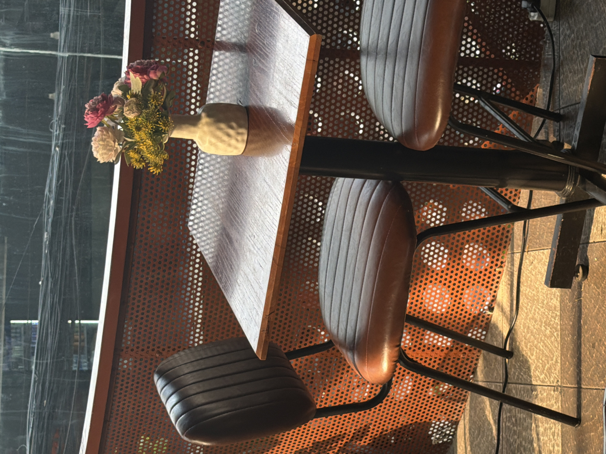 The image displays a static scene within what appears to be a modern cafe or restaurant setting in Đà Nẵng, Vietnam. The central focus is a wooden-topped table with a dark metal base, accompanied by three chairs. The chairs feature dark brown, ribbed, leather-like seats and dark metal frames.
On the table, a light-colored, possibly ceramic or wooden, vase holds a small bouquet of artificial flowers. The bouquet includes pink, cream, and yellow blossoms, along with green foliage.
The background features a prominent orange-brown perforated metal panel, which could serve as a decorative wall or partition. Beyond this panel, a large window or glass wall is visible, reflecting indistinct elements of the surroundings and showing vertical lines, possibly from window coverings or wires.
Strong, natural light suggests it is daytime, likely a sunny morning or afternoon, as evidenced by the distinct shadows cast by the perforated panel onto the table and the tiled or paved floor. These shadows create a pattern of light circles and lines. There is no activity taking place, and no people are visible. No text is discernible within the image.