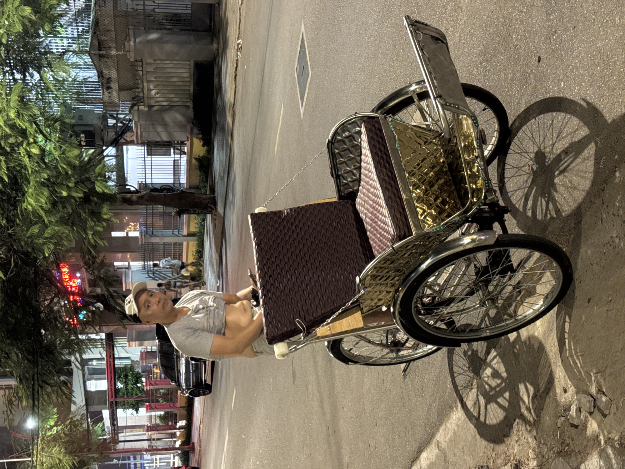 A man in a grey t-shirt and a white baseball cap sits in a pedicab in Nha Trang Ward, Vietnam. The pedicab has a dark brown textured cover on the back and a gold-colored, diamond-patterned material on the sides. The scene appears to be a street at night, with streetlights and illuminated buildings in the background. Trees with lush green foliage are visible to the left, with some colorful neon lights suggesting a commercial area. The man is looking towards the camera with a neutral expression. The street is paved with asphalt, and there is a shadow of the pedicab and its wheels cast on the ground. The lighting suggests it is nighttime, and the weather appears clear. The overall impression is a candid street scene at night in Vietnam.