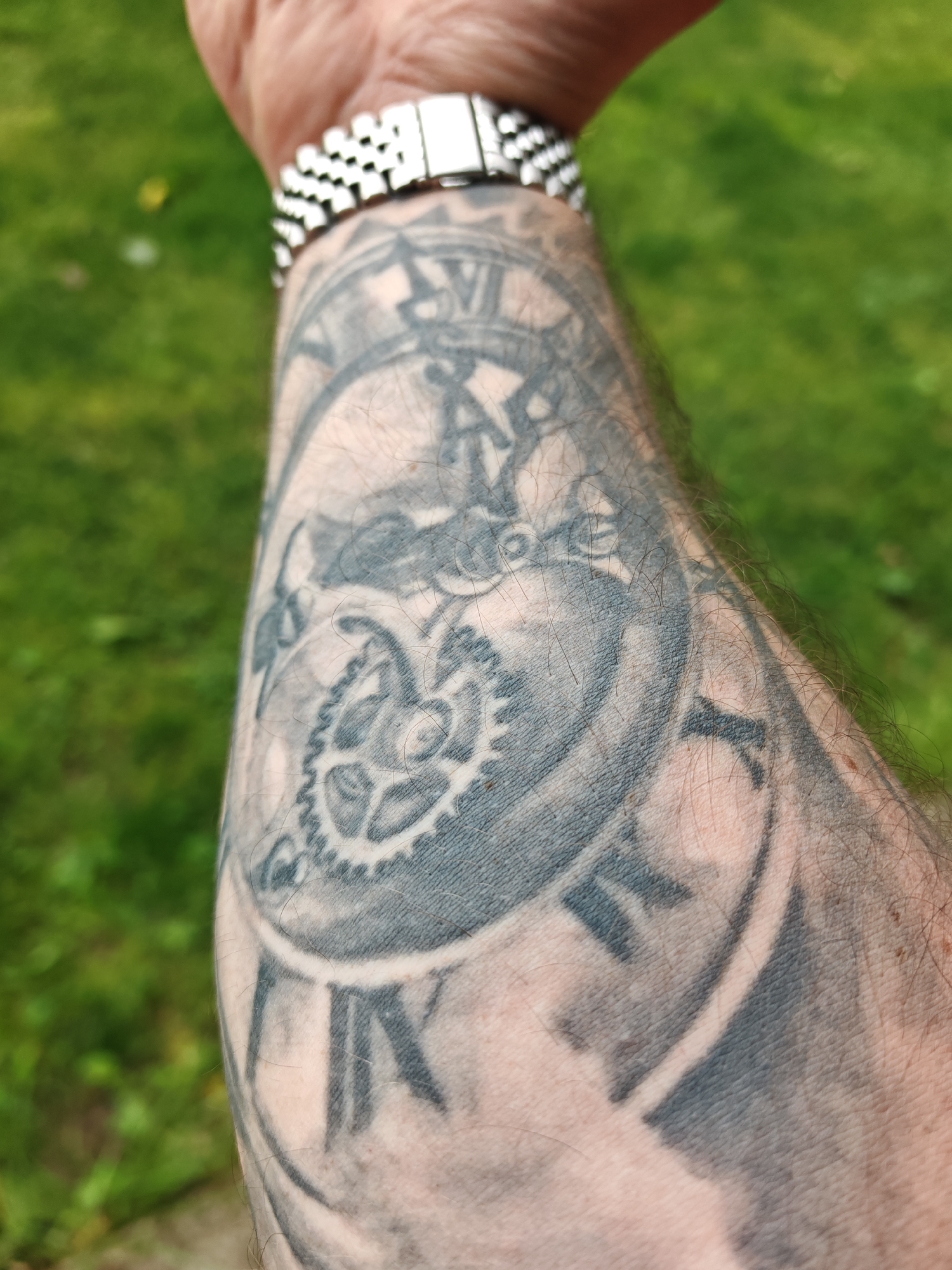 The image depicts a close-up of a person's forearm featuring a tattoo of a clock face with visible gears and Roman numerals. The watch on the wrist is stainless steel. The background is out of focus, showing green grass. There is no indication of Tsarevets, Bulgaria in the picture.