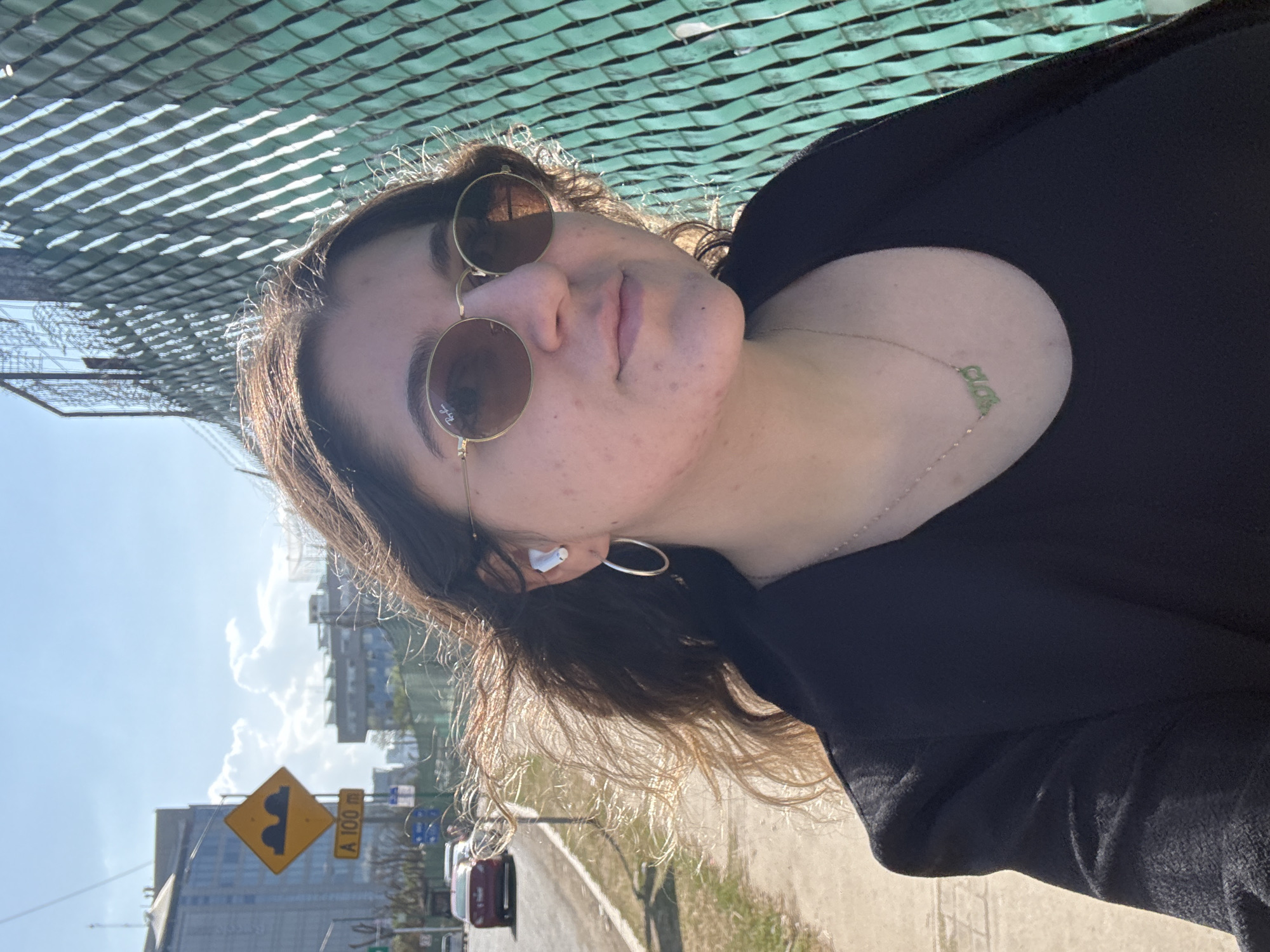 A young woman is depicted in a medium shot, facing slightly right of the camera, likely a selfie. She has long, dark, wavy hair, wind-swept, with highlights from direct sunlight. She wears oval-shaped gold-rimmed sunglasses with dark lenses, a black top, and a black jacket. A silver-colored hoop earring is visible in her left ear, along with a white wireless earbud. A thin necklace with a light green pendant, appearing to be text or numbers, rests on her chest.
She stands on a paved sidewalk next to a road with visible vehicles, including a red sedan. To her right is a green chain-link fence, through which sunlight casts distinct shadows. In the background, under a clear blue sky with white clouds, urban buildings are visible. A yellow diamond-shaped road sign depicting two speed bumps, with a rectangular sign below it reading "A 100 m," identifies the location as Mexico City, Mexico.