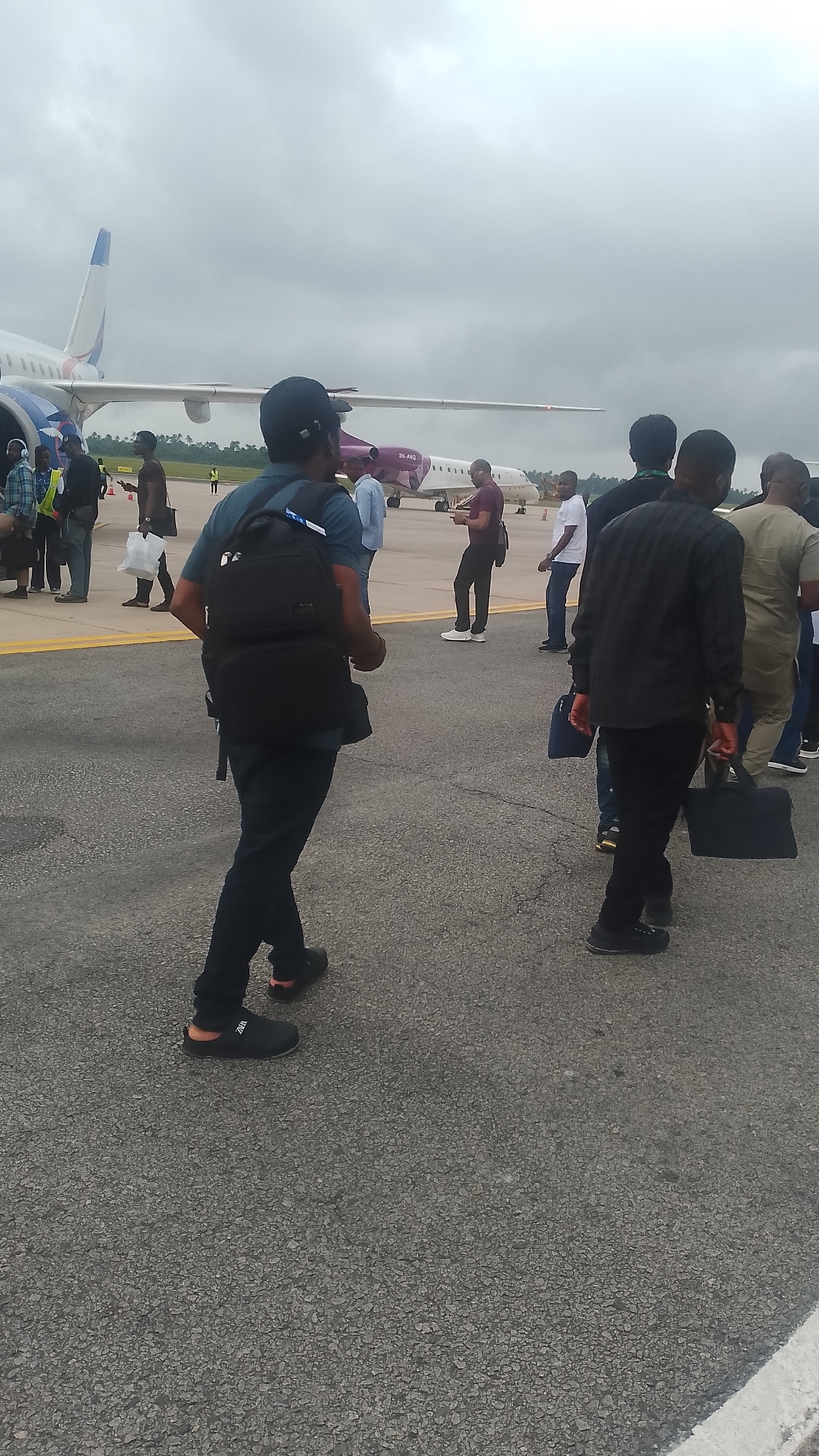 An overcast day at the Orerokpe Airport, Nigeria. Several individuals are gathered on the tarmac, walking towards or standing near two airplanes. In the foreground, a person wearing a black backpack and dark pants walks away from the camera. To the left, a group of people stand near an airplane, some in casual attire, one in a high-visibility vest. Further to the right, another group of individuals, mostly men dressed in dark clothing, walk in the same direction as the person in the foreground, some carrying bags. The environment is paved with asphalt, and the sky is heavily clouded.