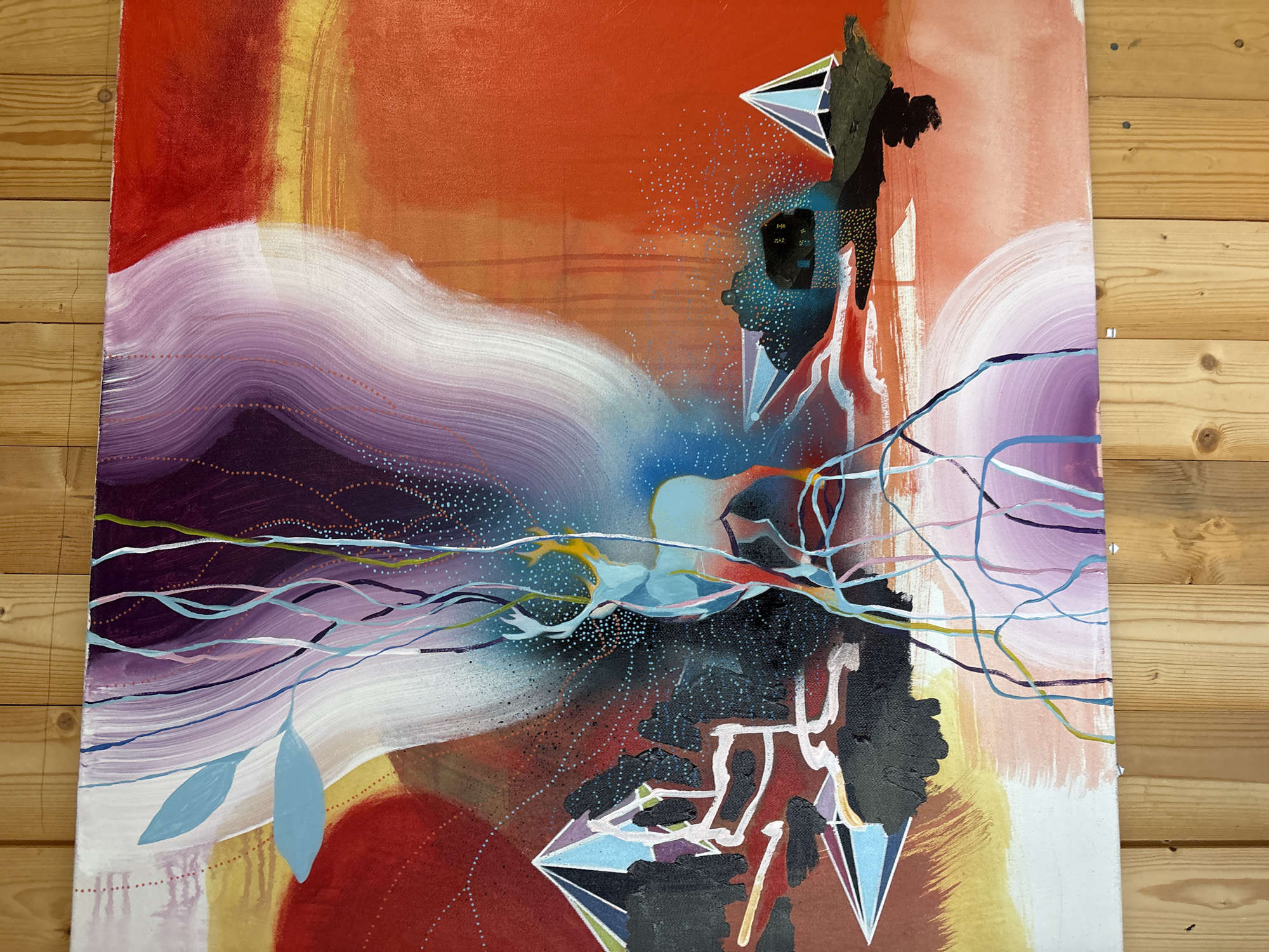 This is an oil painting on canvas, leaning against a wooden wall. The artwork depicts a surreal landscape with a stylized deer as the central subject. The deer is light blue and outlined in thin, flowing lines of purple, blue, and yellow. It is positioned in the center of the canvas, emerging from a cluster of dark, angular shapes that resemble trees or rocks. Above the deer, thin, colorful lines cascade down, resembling abstract vines or streams of energy.
The background features bold, abstract washes of color, primarily in shades of red, orange, yellow, white, and purple. Large, curved brushstrokes create a sense of movement and depth. Scattered across the canvas are small, dotted patterns, some resembling stars or a constellation. Geometric shapes, resembling faceted crystals or mountains, are present on the left and right sides of the composition. On the right side, a small, dark structure, possibly a house, is nestled within a dark, textured area.
The painting is framed by natural wood planks, which are visible at the top and bottom of the image. The wood has a light, grainy texture and is held in place by visible nails. The overall impression is one of a vibrant and imaginative scene, blending natural elements with abstract and fantastical imagery.
There is no discernible location in the city of Gryllefjorden, Norway. The image is of an artwork, not a photograph of a real-world location.