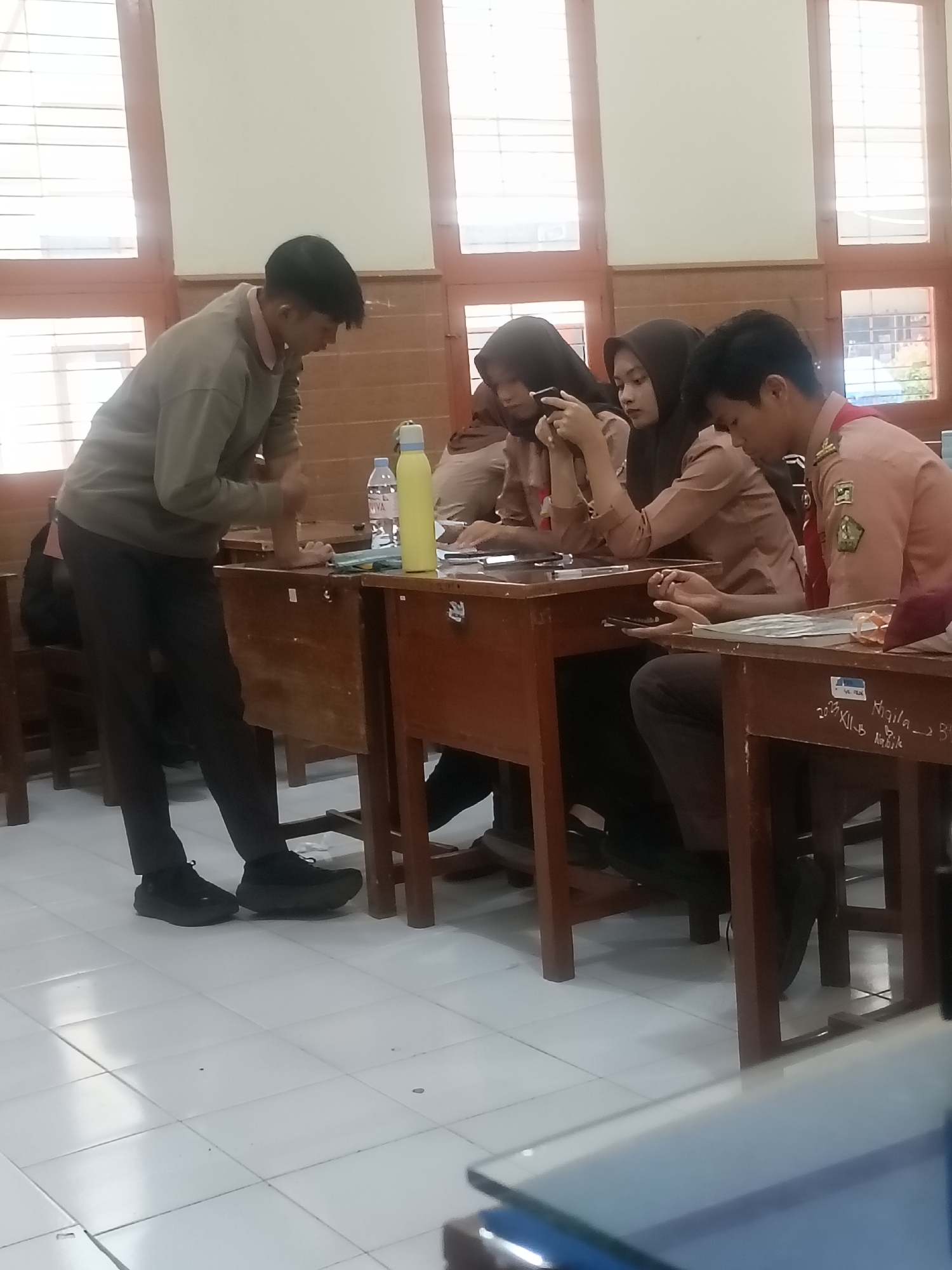 The image depicts a classroom scene in Kediri City, Indonesia. Four individuals are present, three seated at desks and one standing. The seated individuals appear to be students wearing uniforms, with two wearing headscarves. One student is looking at a smartphone, and another is holding a smartphone. The standing individual, also possibly a student, is wearing a light brown sweater and dark pants. A yellow water bottle and a bottle of "IVANA" water are on one of the desks, along with papers and a tablet. The room has wooden desks and chairs, with windows covered by blinds in the background. The floor is tiled. The overall atmosphere suggests an educational setting, potentially during a lesson or break.