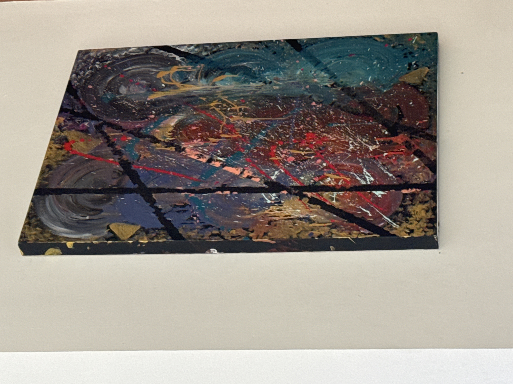 A square abstract painting hangs on a white wall. The painting is predominantly dark, with swirling patterns of blue, black, brown, red, and gold paint. Black lines cut across the painting, forming a grid-like structure. The painting is likely in an interior space, but its specific location in Denpasar, Indonesia, is not discernible.