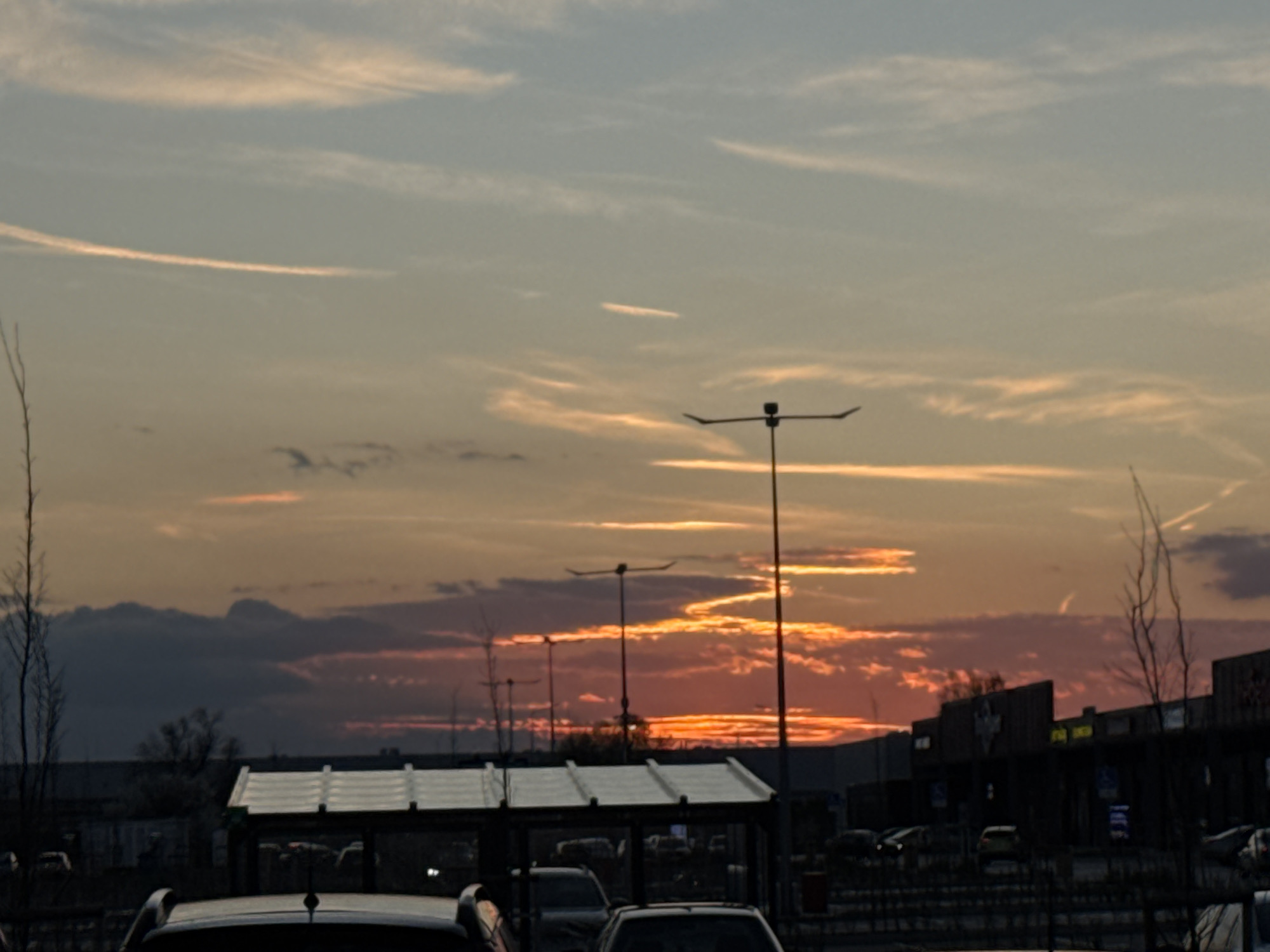 The image shows a sunset over a parking lot in Sofia, Bulgaria. In the foreground, there are several parked cars and a bus stop. Beyond that are streetlights and a building with signage. The sky is filled with orange and purple clouds, indicating either a sunrise or a sunset. Vapor trails are visible. There are few trees in the scene.