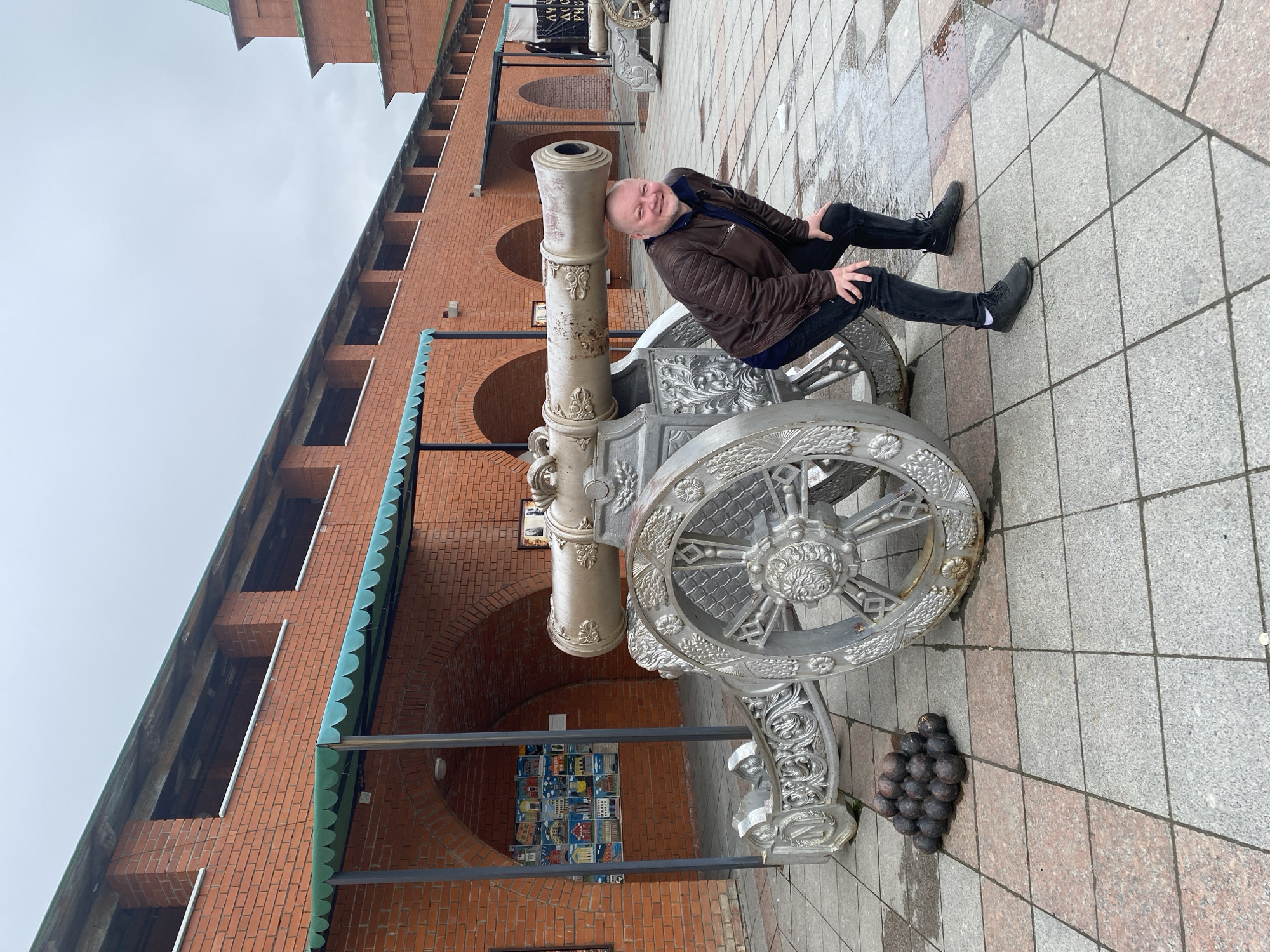 The image, when correctly oriented, captures a man sitting on a large, elaborately decorated cannon in an outdoor setting, likely a historical or cultural area in Yoshkar-Ola, Russia.
The subject is a bald man with a fair complexion, dressed in a brown leather jacket, a dark blue shirt, and dark trousers. He is smiling warmly at the camera, with his hands resting casually on his knees, suggesting he is posing for a photograph.
The prominent object is the impressive cannon, featuring intricate decorative carvings on its barrel, carriage, and large, spoked wheels. It appears to be made of metal with an aged, metallic silver-grey finish. Adjacent to the cannon's wheels, a neat pile of black, spherical cannonballs is visible on the ground.
In the background, a substantial building constructed from red bricks forms a long facade with numerous arched openings. A distinctive green, scalloped trim adorns parts of the building's lower roofline or awning. The ground is paved with square tiles in a pattern of grey and reddish-brown hues. Under one of the arches, a colorful display of small images, possibly artwork or photographs, is arranged in a grid.
The weather appears to be overcast, with a grey sky, and sections of the pavement look damp, suggesting recent rain. The man's cheerful expression conveys a relaxed and enjoyable atmosphere during his visit. Some Cyrillic text is present on signs in the background, but it is too indistinct to be clearly read.