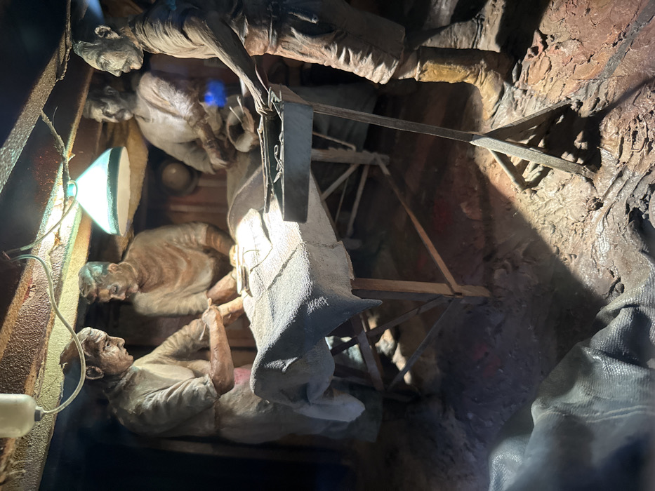 The scene is a diorama depicting a surgical operation, located at the Imperial War Museum, London, United Kingdom (latitude: 51.497, longitude: -0.176). The scene is lit by a single overhead lamp. There are four figures in the diorama, all dressed in period clothing. Two figures are positioned on either side of a surgical table, operating on a figure lying prone on the table. The fourth figure is standing to the right of the table, observing the operation. The table is covered with a white sheet and contains a metal tray with surgical instruments. There are various objects scattered around the floor, including a single cloth bag and wooden floor boards. The diorama is surrounded by a dark, wooden frame.