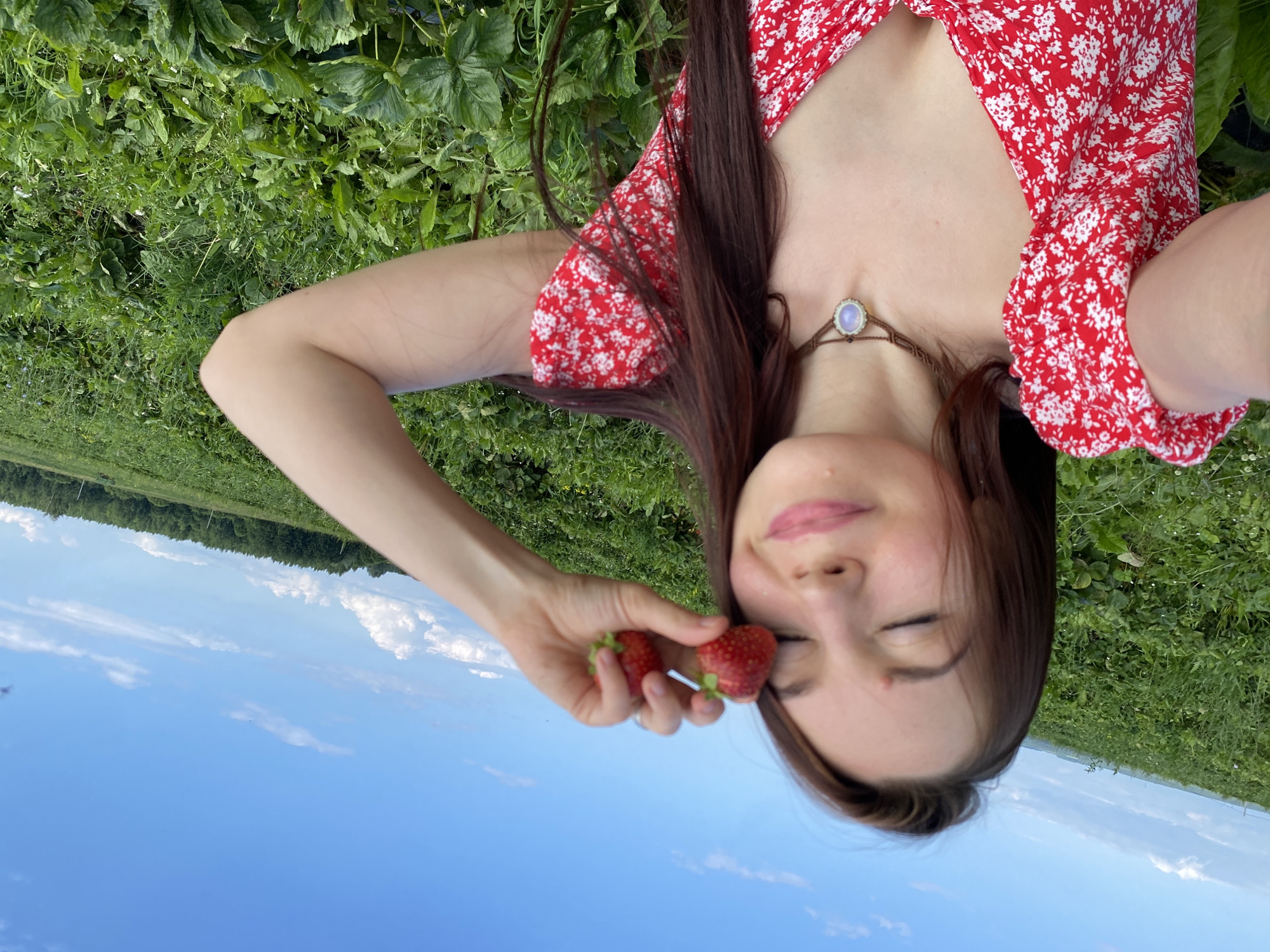 A young woman with long brown hair lies on her back in a field of green plants, possibly a strawberry patch, with the bright blue sky and some white clouds above. She is wearing a red and white floral print dress. Her eyes are closed, and she has a gentle smile on her face. In her right hand, she holds two ripe strawberries near her cheek. The image has a relaxed and peaceful atmosphere, suggesting a warm and sunny day, likely in the summer. The presence of the field and the sky implies an outdoor setting, possibly a rural area. There is no visible text in the image. The location context is Foteni, Russia.