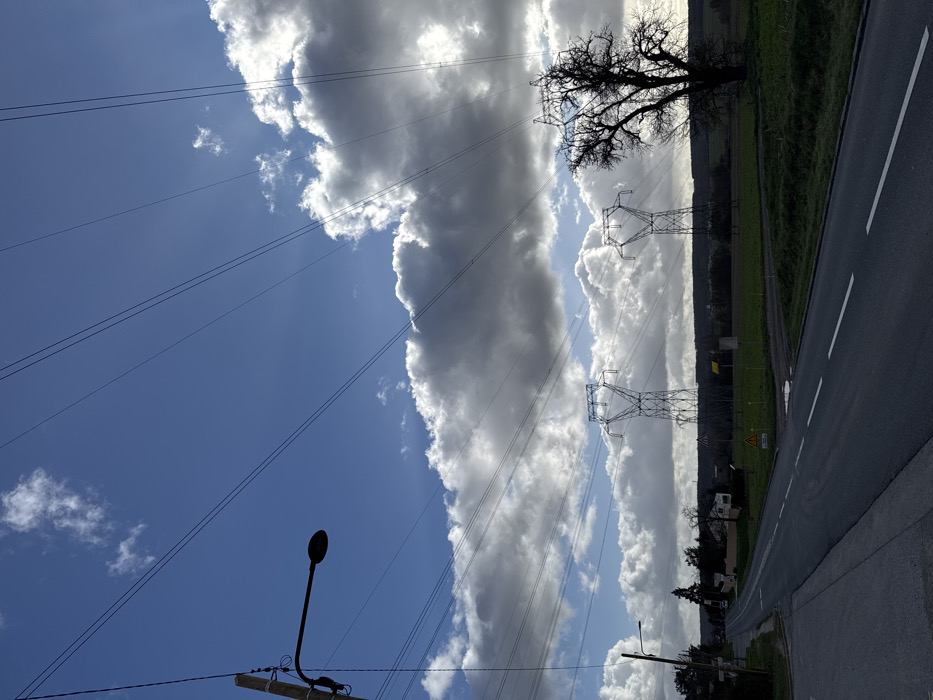 This image depicts a rural or semi-rural scene in Marcoussis, France, under a partially cloudy blue sky. Overhead, numerous power lines crisscross the upper portion of the frame. On the far left, a wooden utility pole supports a black, downward-curving streetlamp. Below it, a paved two-lane road with a solid white edge line and a broken white center line curves from the lower-left foreground, where a sidewalk is visible, towards the center and recedes into the distance on the left.
To the right of the road, a green grassy field extends towards the horizon. In this field, two large, grey metal lattice-type electrical transmission pylons are positioned in the midground, carrying multiple power lines, some of which are visible against the sky. A prominent, large, bare deciduous tree with dark, intricate branches stands on the far right, silhouetted against the bright clouds. A yellow triangular warning sign is visible in the grassy field near the road.
In the background along the road, several residential structures and clusters of trees are visible, indicating a populated area. The sky features bright white cumulus clouds, with strong sunlight illuminating the scene from the upper-right, creating sunbeams through the cloud cover. No individuals or vehicles are present or in motion within the frame.