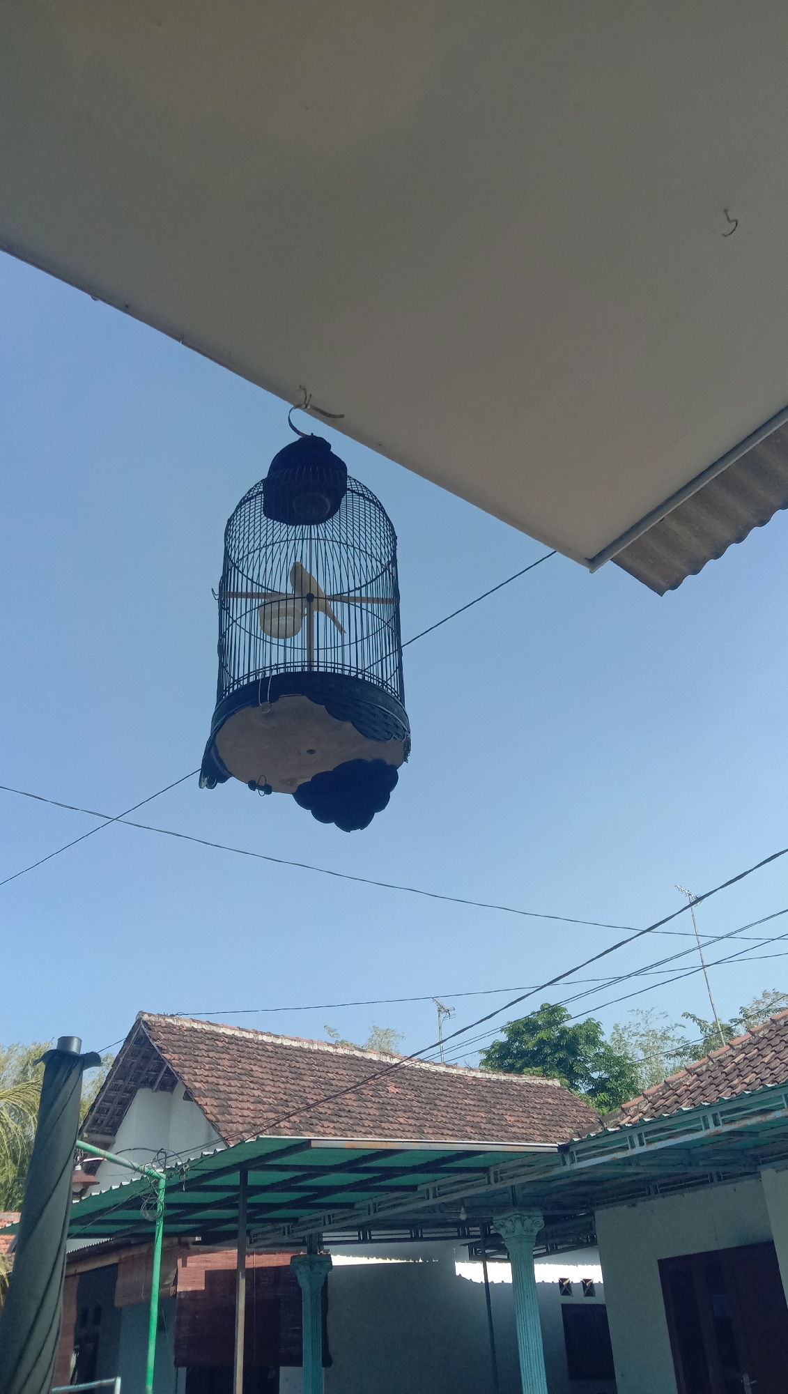The image depicts a birdcage hanging outdoors in what appears to be a residential area. The birdcage is black and cylindrical, with a decorative base and a perch inside. It is suspended by a hook from a light-colored overhang. The background is dominated by a clear blue sky. Below the overhang, the roof of a building with terracotta tiles is visible, along with supporting structures that suggest a covered patio or porch. In the distance, trees and more rooftops can be seen. The scene suggests a warm, sunny day. The location is identified as Sidogiri, Indonesia.
