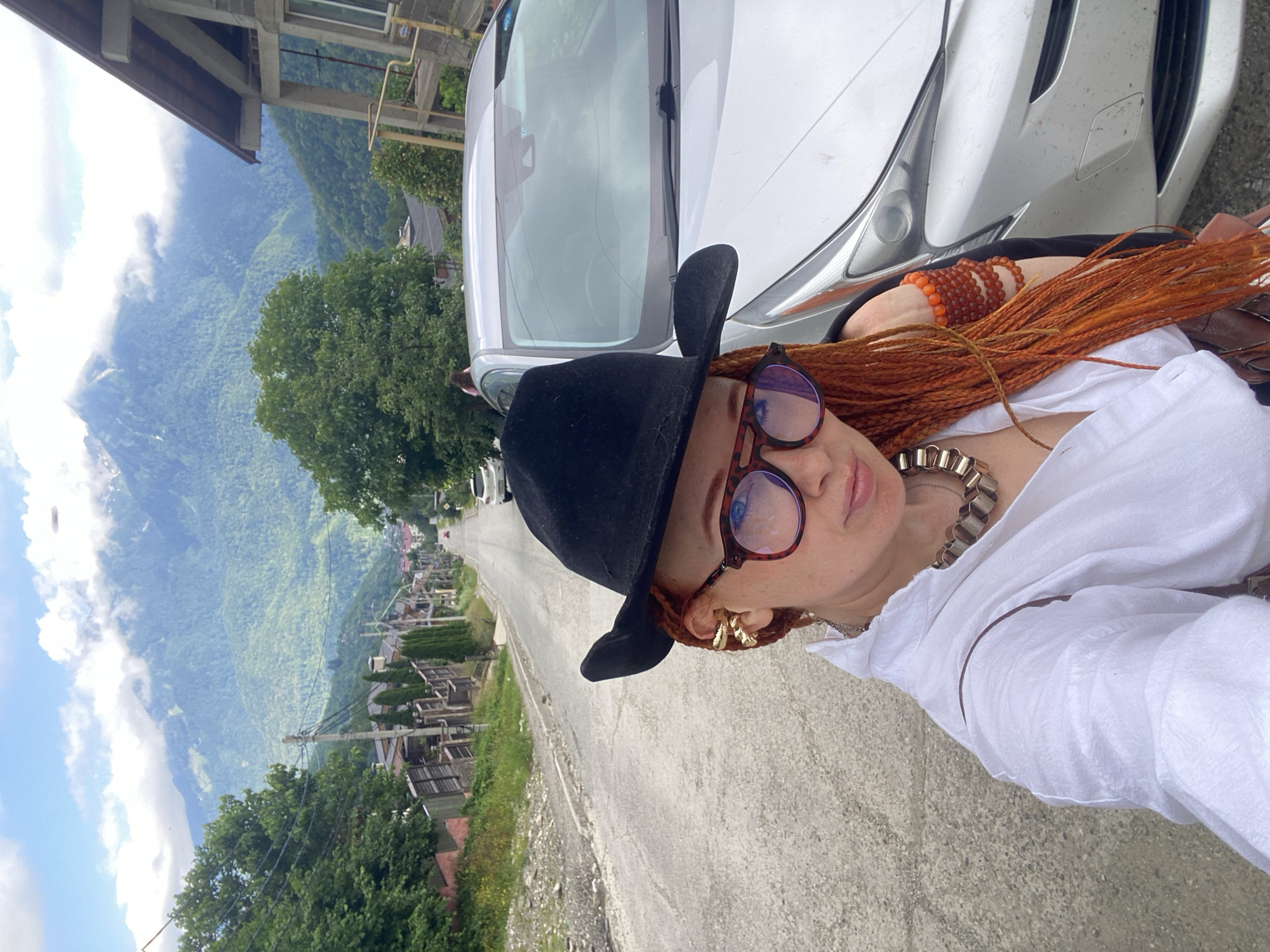 The image shows a woman with auburn braided hair and a black hat, wearing large-rimmed glasses and a white shirt. She is wearing multiple beaded bracelets on her wrist and a distinctive metallic necklace. She is taking a selfie outdoors.
The background features a steep, winding road lined with small houses and dense green trees, suggesting a mountainous or hilly terrain. In the distance, lush, green mountains rise, partially shrouded in clouds, indicating a misty or overcast day. A silver car is parked on the right side of the frame, partially obscuring the woman.
The scene appears to be in Krasnaya Polyana, Russia, a location known for its mountainous landscape and ski resorts, which aligns with the visual cues of the image. The lighting suggests it is daytime, with diffused light due to the cloud cover. The woman's pose and direct gaze into the camera indicate she is intentionally capturing a moment. There is no visible text in the image.