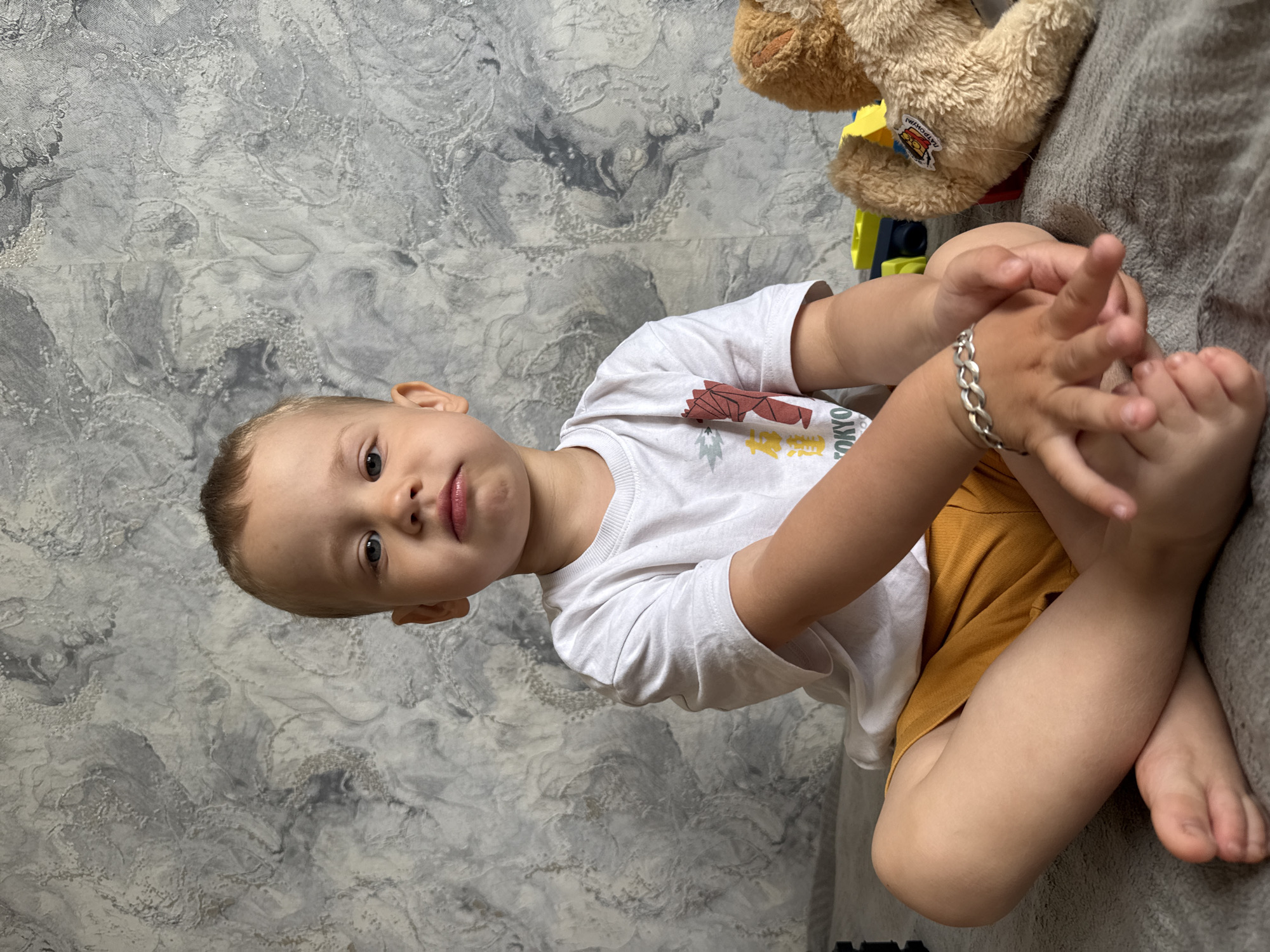 A toddler with fair skin and blue eyes is sitting cross-legged on a gray surface, likely a couch or bed, with a gray and white textured wall in the background. The toddler is wearing a white t-shirt with a graphic print and mustard-colored shorts. The child has a silver bracelet on their left wrist and is holding their toes with both hands, gazing directly at the camera with a slightly puckered expression. A partially visible brown teddy bear is situated in the upper right corner of the image. Plastic blocks, yellow and blue, are also visible near the bear. The location is unknown and there is no confirmation that the location is in Rusanovo, Russia.