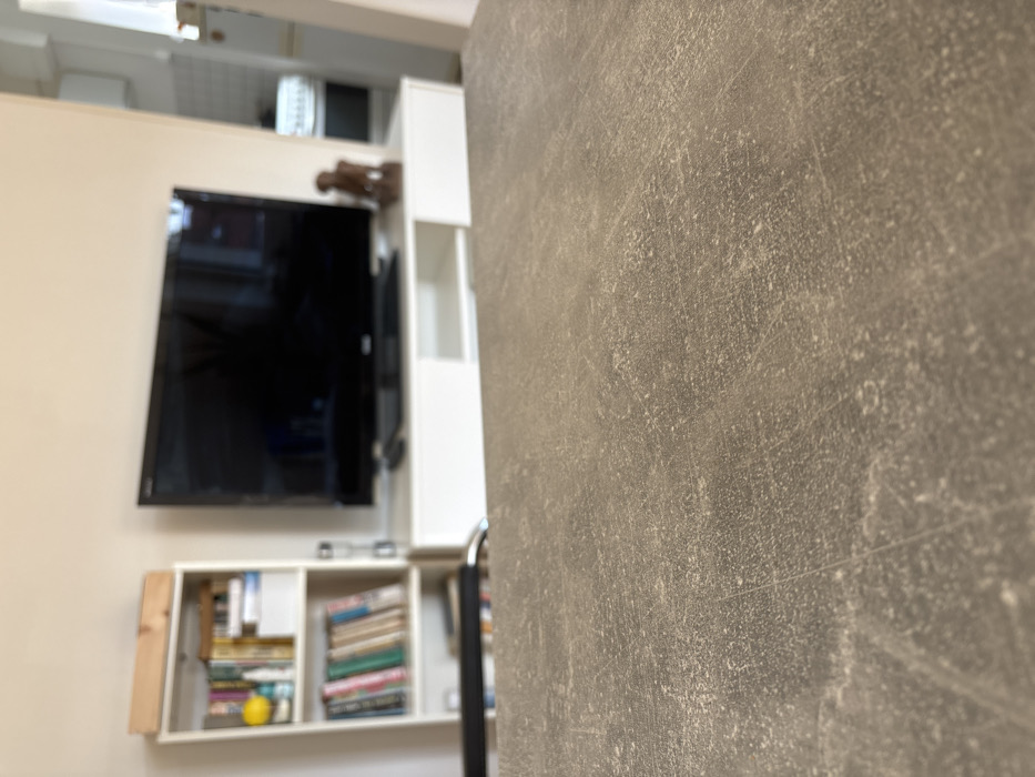 The image depicts an interior residential space, likely a living room, in Germering, Germany. The foreground features a close-up view of a textured, matte grey-brown surface, possibly a tabletop or countertop, which occupies the lower half of the frame and is in focus.
The mid-ground and background are blurred. Centrally positioned is a large, black flat-screen television with its screen off, resting on a white, low-profile media console. A small, dark object, resembling a remote control, is visible on the console to the left of the TV. To the right of the television on the console, a dark brown, abstract statuette or sculpture of a humanoid figure is present.
To the left of the television setup, a white, vertical bookshelf contains multiple rows of books, a yellow spherical object, and other indistinguishable items. A light-colored wooden box rests horizontally on top of the bookshelf.
To the right of the media console, an architectural opening reveals a glimpse into an adjacent room, possibly a kitchen, where a white appliance or counter is partially visible. The walls throughout the visible area are painted a uniform, light, neutral color, consistent with beige or off-white. No individuals are present, and no active interactions or specific actions are depicted within the scene. The lighting suggests an ambient, indoor environment.