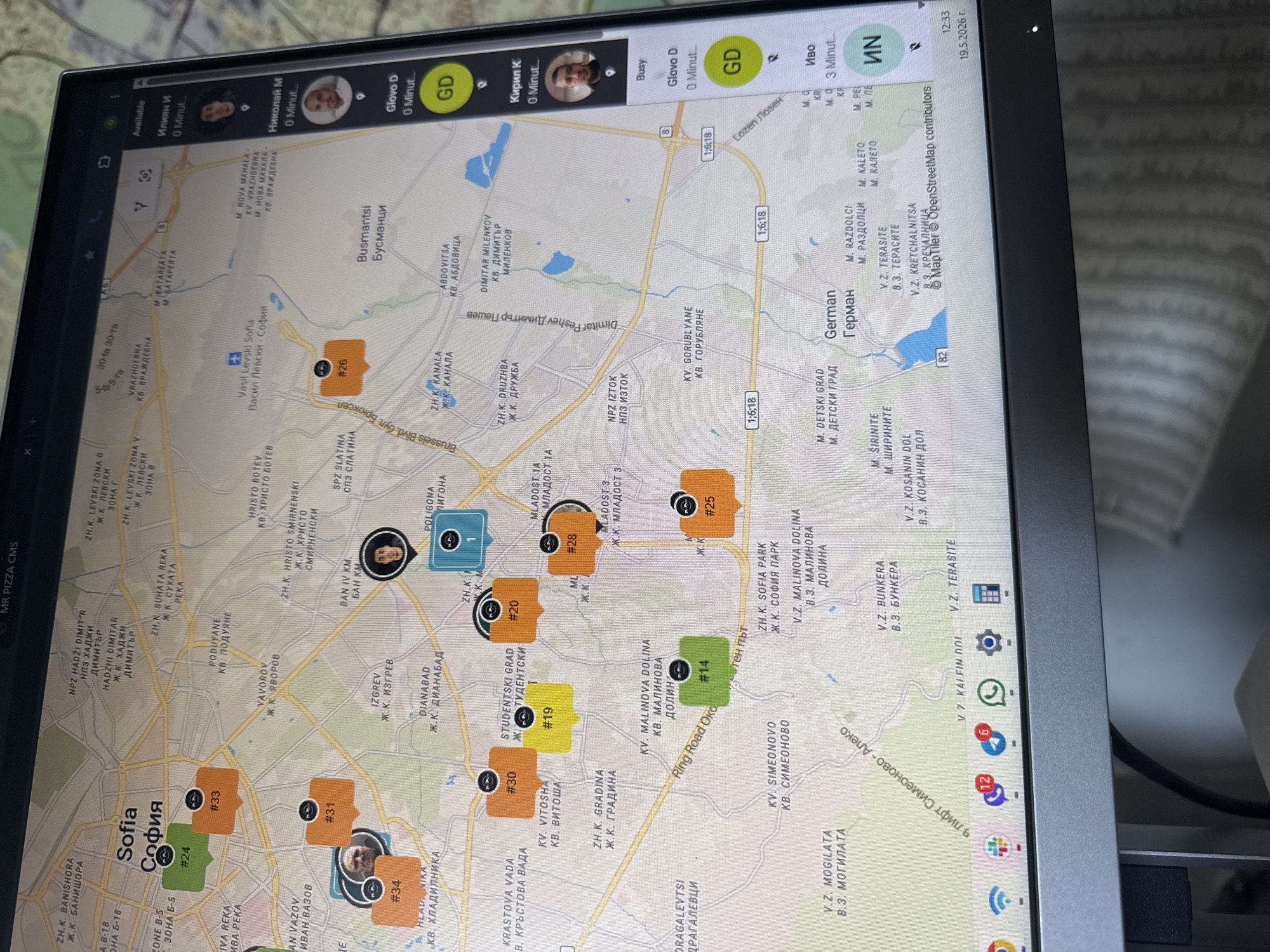 The image shows a computer screen displaying a map of Sofia, Bulgaria, along with user interface elements. The map is densely populated with location markers, numbered and colored, indicating different areas of interest. Place names like "Sofia", "Vitosha", "Malinova Dolina", and "Studentski Grad" are visible. The interface shows user avatars with names like "Илиян И", "Николай М", "Кирил К", and "Иво". There is also a date: "19.5.2026." Icons and notifications are visible at the bottom of the screen. The environment includes a desk where the screen sits.