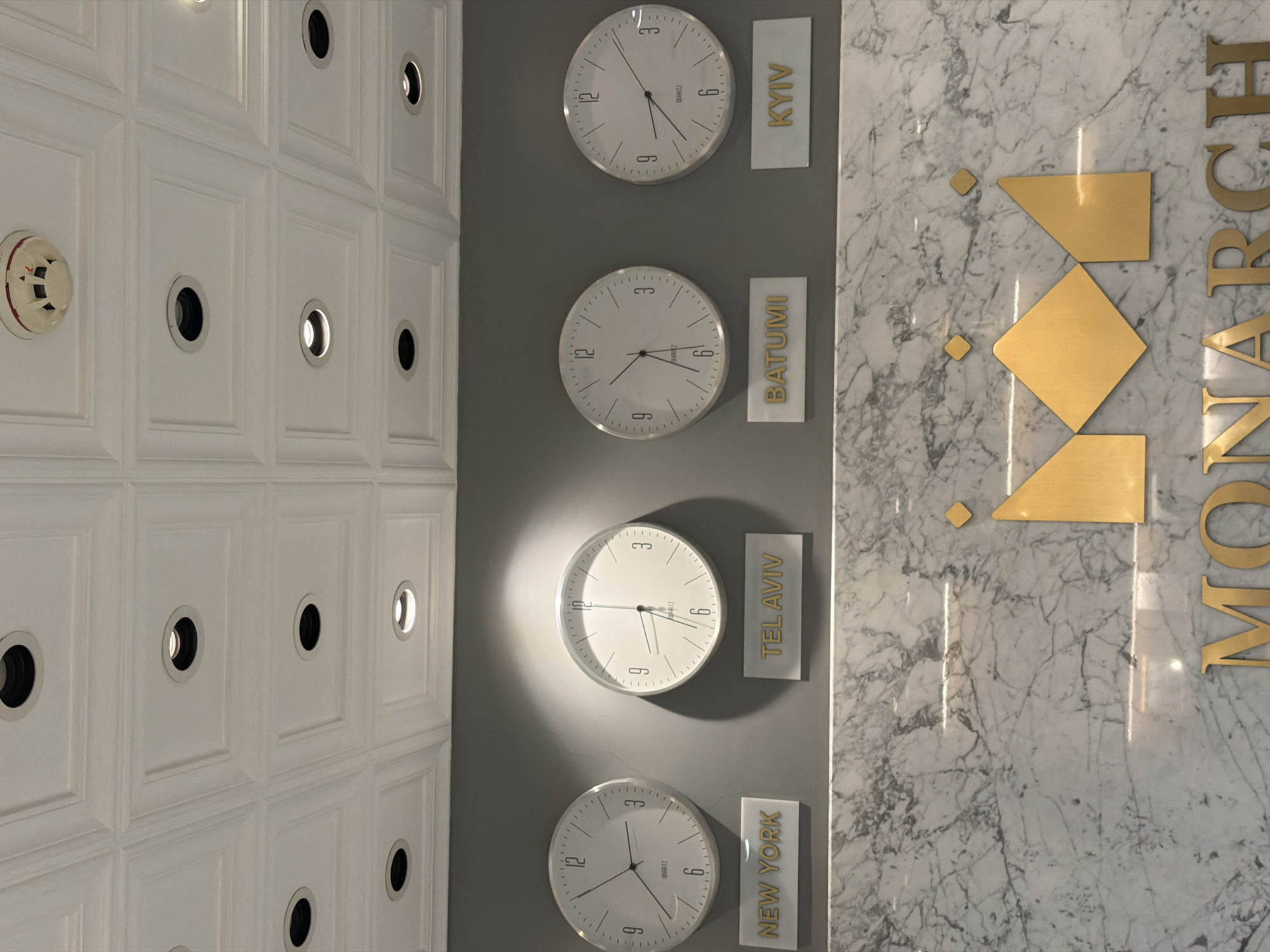 The image shows a wall in what appears to be a building, potentially a hotel or office space. The wall is divided into three sections. On the left is a white paneled wall with inset circular lights or possibly cameras, and a fire alarm. The middle section features a gray wall with four clocks displaying different times and labeled with city names: Kyiv, Batumi, Tel Aviv, and New York. The right section is a white marble wall with gold-colored geometric shapes and parts of the word "MONARCH" vertically written in gold. The clocks likely indicate the time zones of those cities in relation to the location of the Monarch hotel. The arrangement suggests a global or international theme, commonly found in hotel lobbies or business environments. There are no people visible.