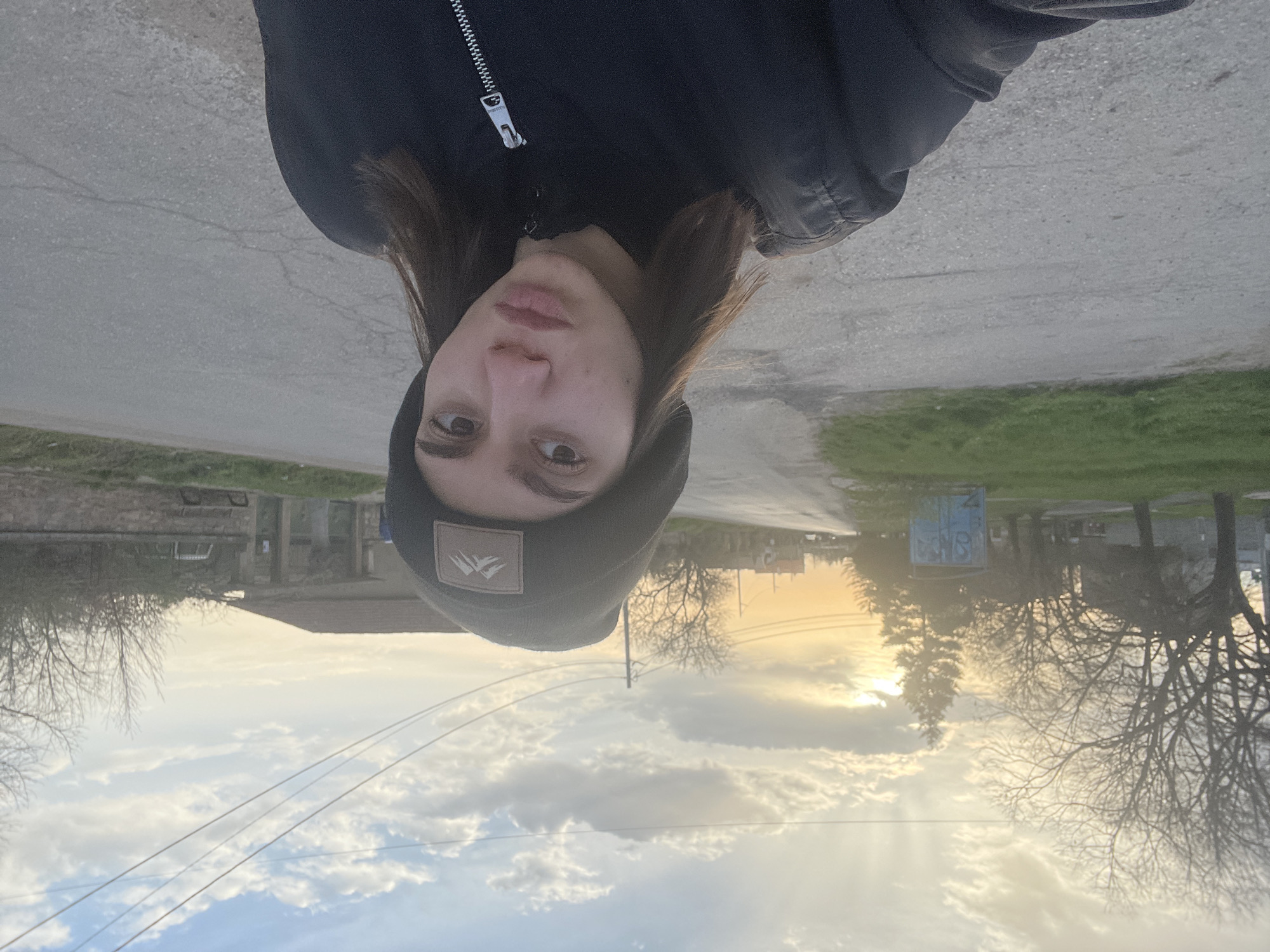 A selfie-style photograph shows a young individual with fair skin, brown hair, and a black beanie with a logo. They are wearing a dark-colored jacket. The photo appears to be taken outside, with a cloudy sky and the setting sun visible in the background. The surrounding environment suggests a rural or suburban area, with leafless trees, buildings, and power lines in the distance. The photograph is oriented upside down. Based on available information, this photo may have been taken in Aprilovo, Bulgaria.