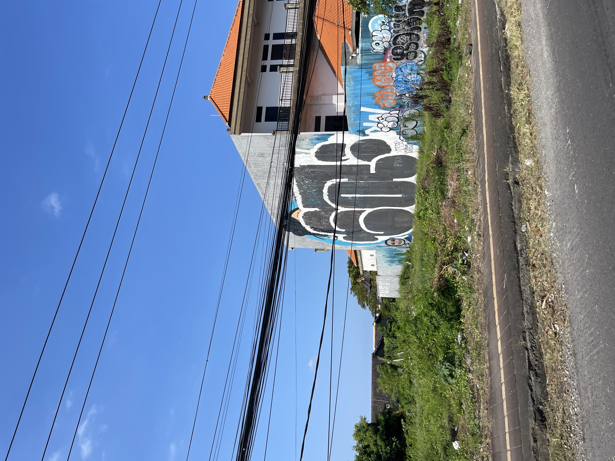 In Kuta, Indonesia, a building with a red-tiled roof and white exterior is visible. The building features multiple windows and a balcony on the upper level. The wall of a lower section of the building is covered in graffiti, with large black and white lettering dominating the design. Smaller, more colorful graffiti tags are also present on the lower portions of the wall. Adjacent to the building is an overgrown patch of green vegetation. Several black electrical wires stretch across the bright blue sky, creating a layered effect. A paved road and sidewalk are in the foreground, with the sidewalk featuring tactile paving. The scene appears to be an outdoor environment on a sunny day.